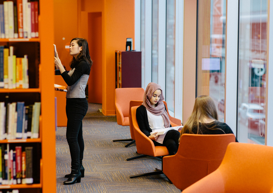 Three women are in a modern library; one stands browsing bookshelves, while two others sit in orange chairs by large windows, reading quietly.