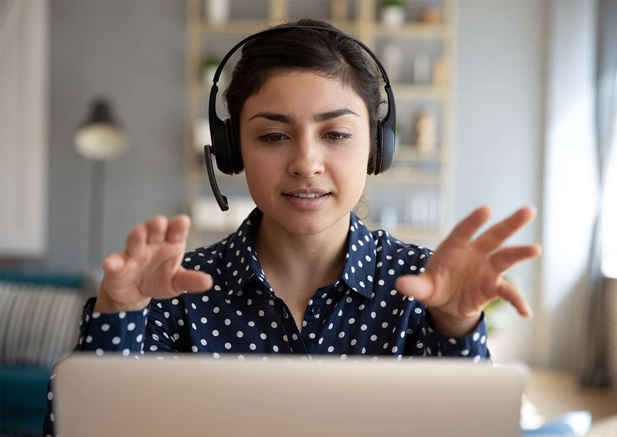 Woman wearing a headset sits in front of a laptop, gesturing with her hands while participating in a video call in a home office setting, adapting during a crisis with confidence and effective communication.