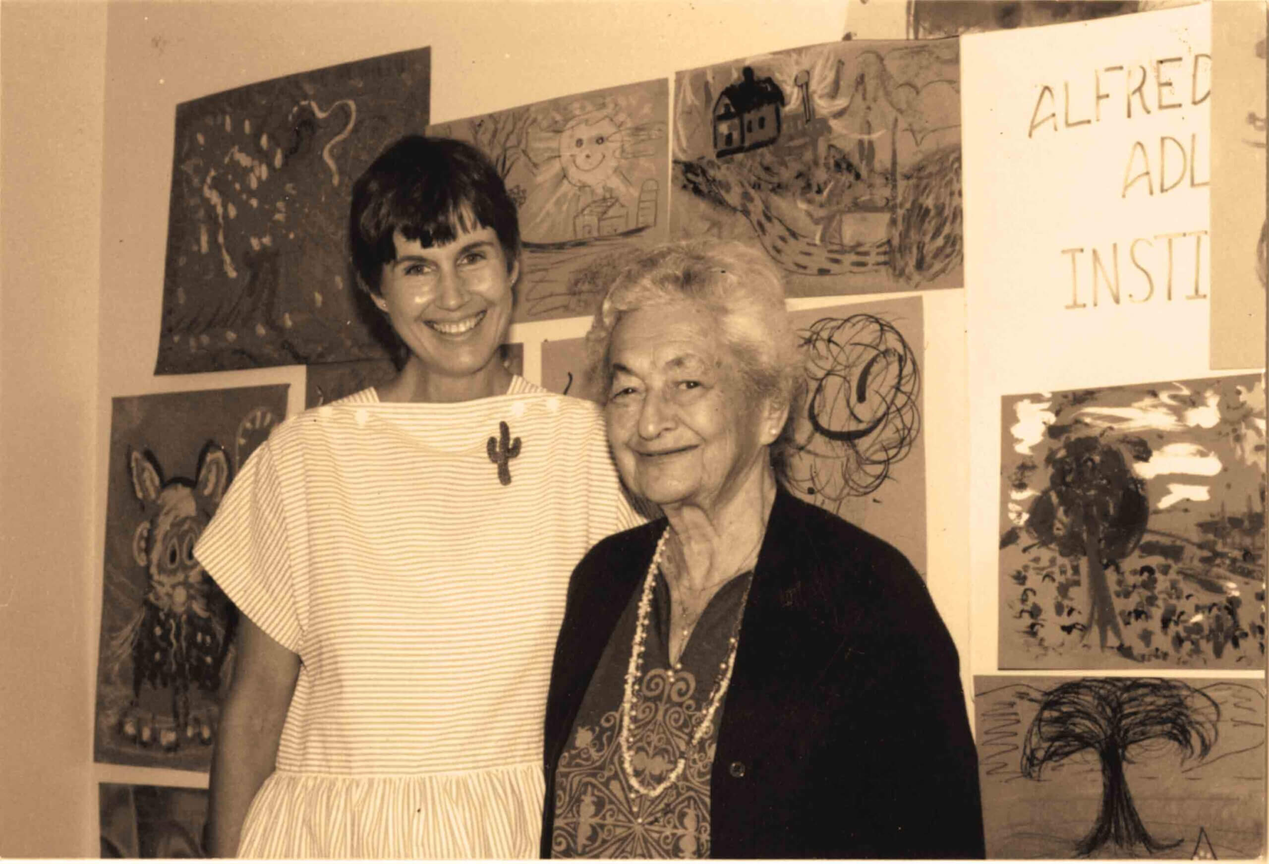 Two women stand in front of a wall displaying children's artwork and a partial sign reading "Alfred Adl... Instit...".