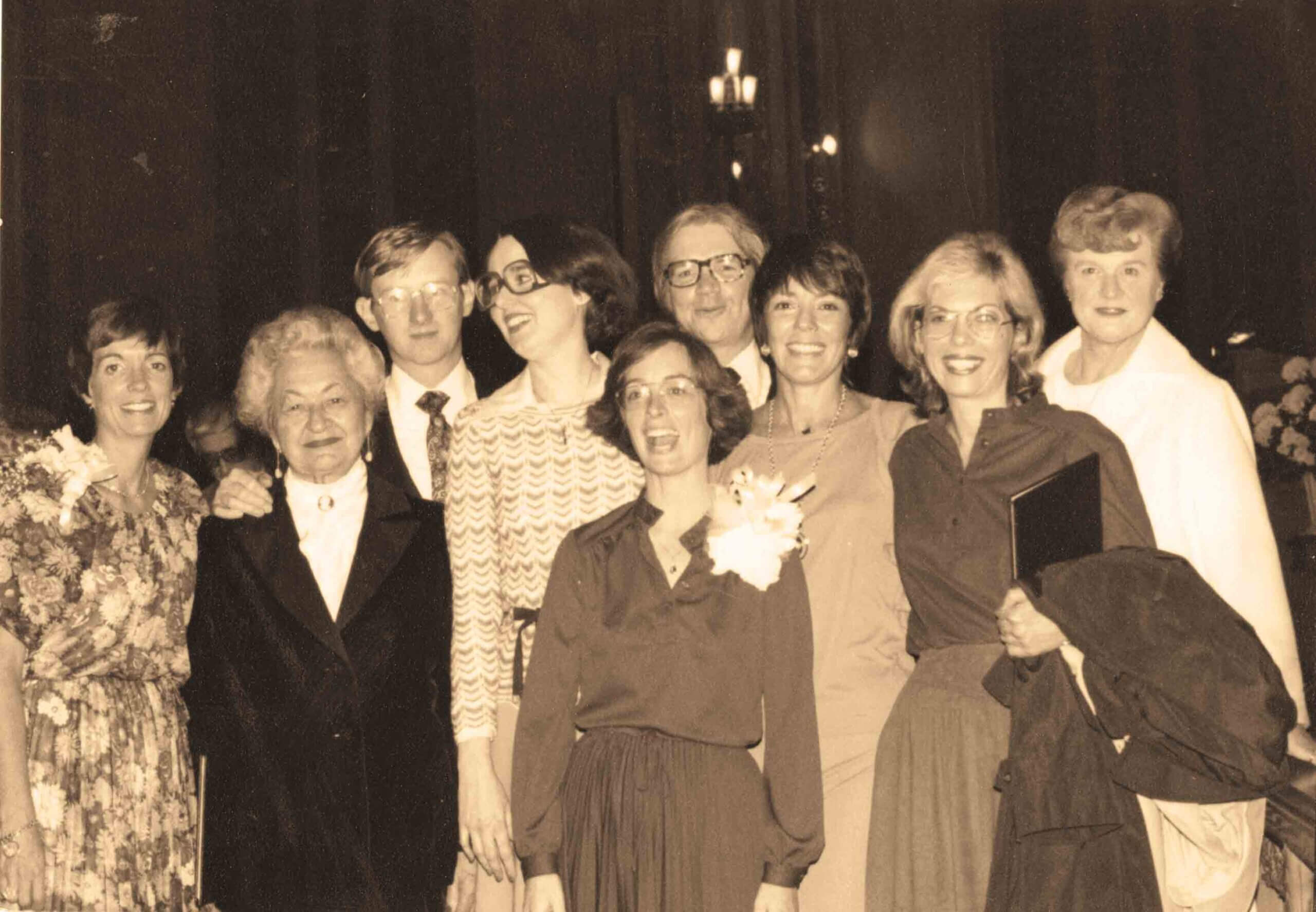 A group of nine adults posing together indoors, dressed in formal and semi-formal attire, with some smiling at the camera. The image is in sepia tone.