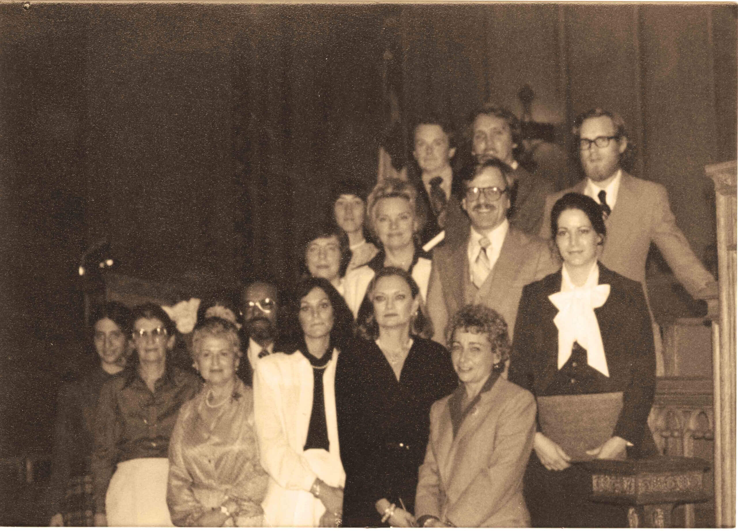 A sepia-toned photo of thirteen people, men and women, posed in formal attire inside a church or formal setting, arranged in two rows with some standing and others seated.