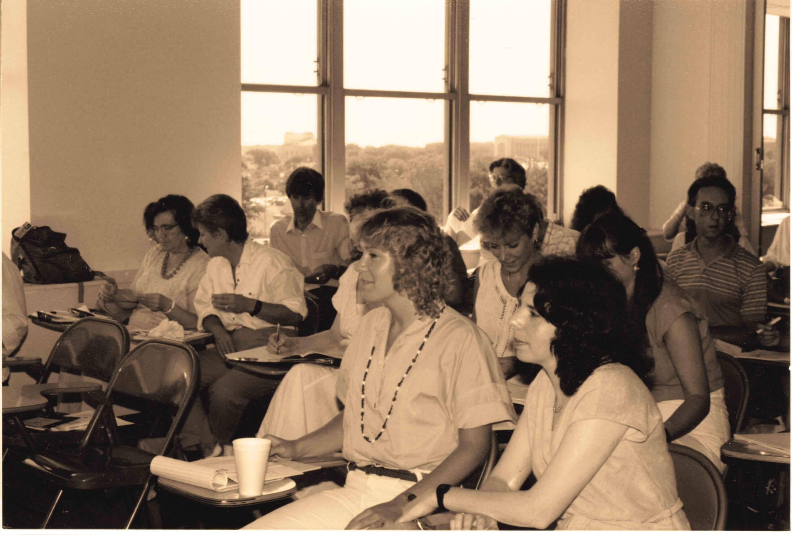 A group of adults sits in a classroom, taking notes and listening to a presentation. Large windows in the background let in natural light.
