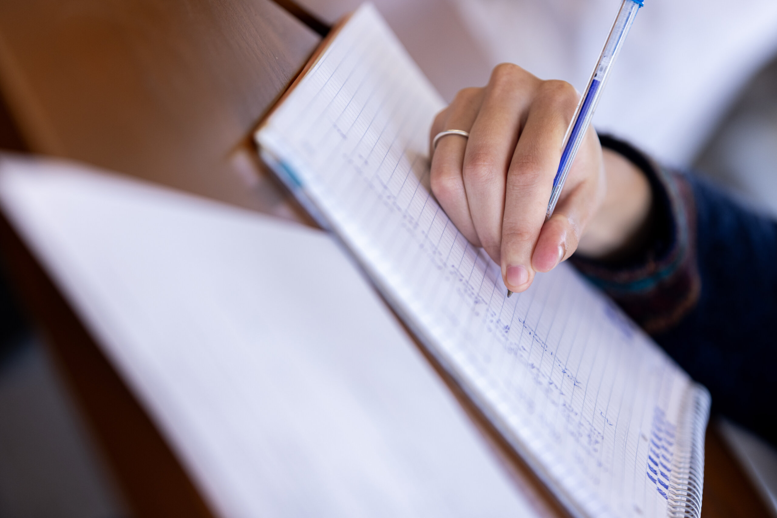A person writes with a pen in a notebook on a desk, with sheets of paper nearby.