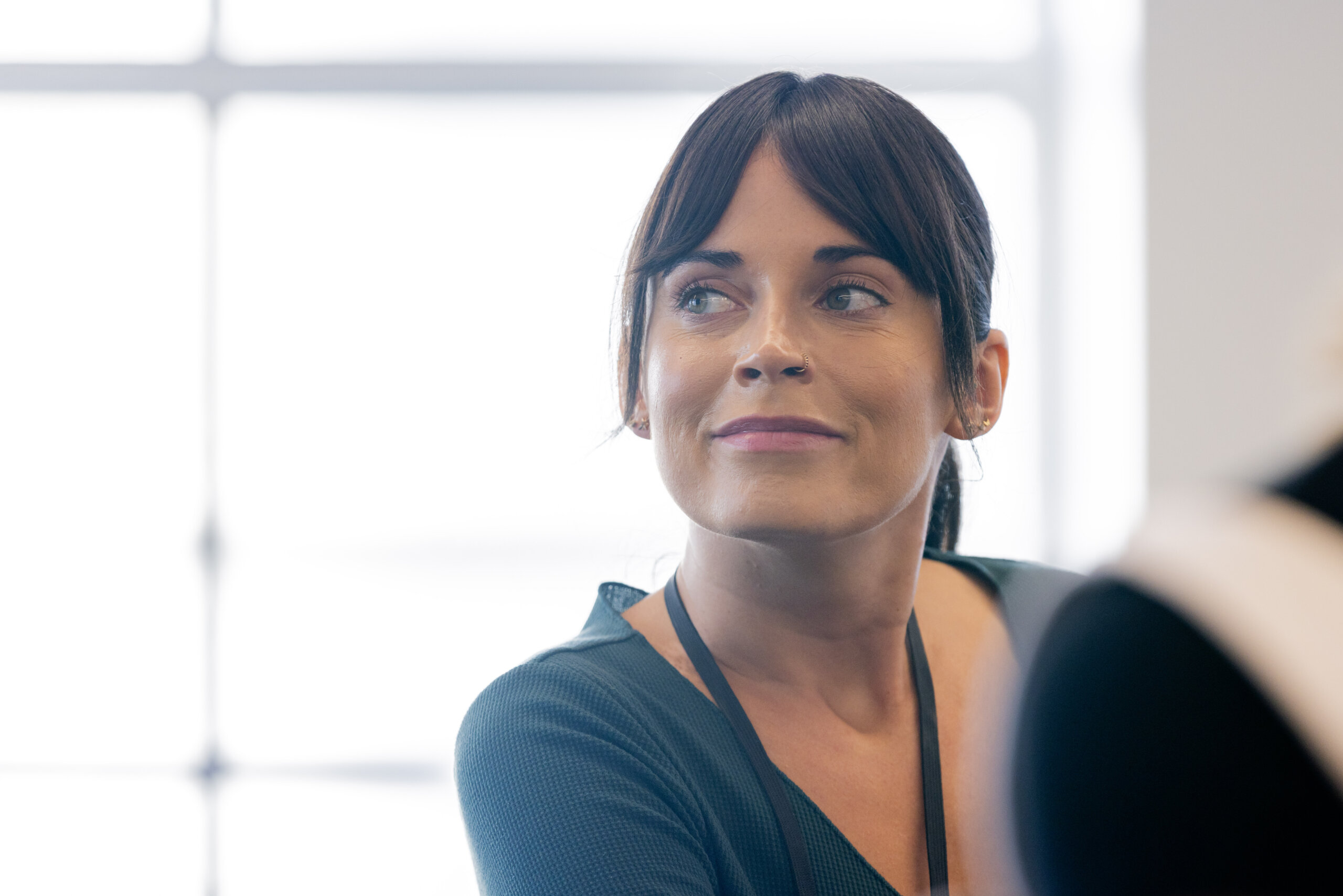 A woman with dark hair and bangs, wearing a green top and lanyard, looks slightly to the side in a brightly lit indoor setting.