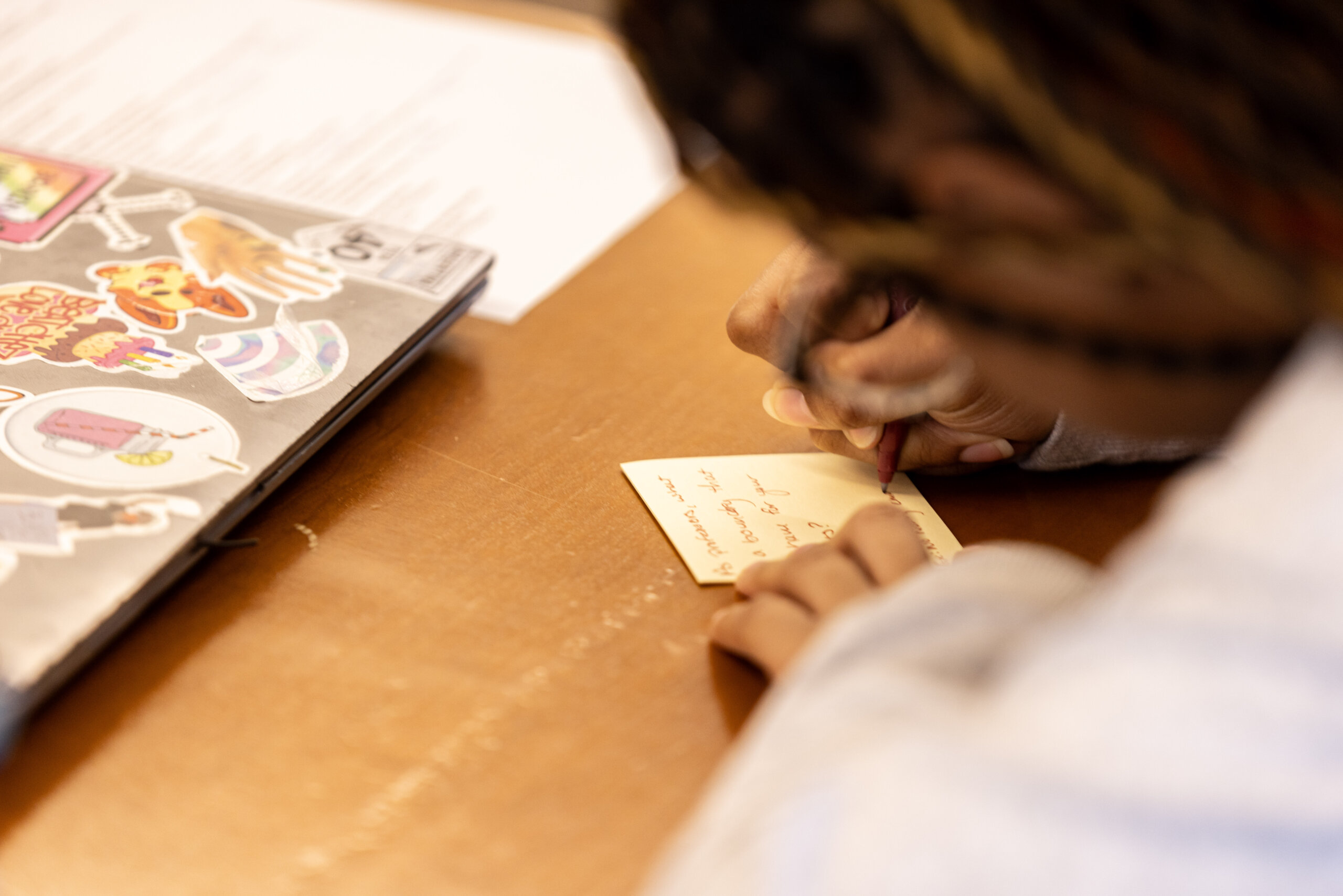 A person writes on a small piece of paper at a desk next to a laptop decorated with colorful food-themed stickers.