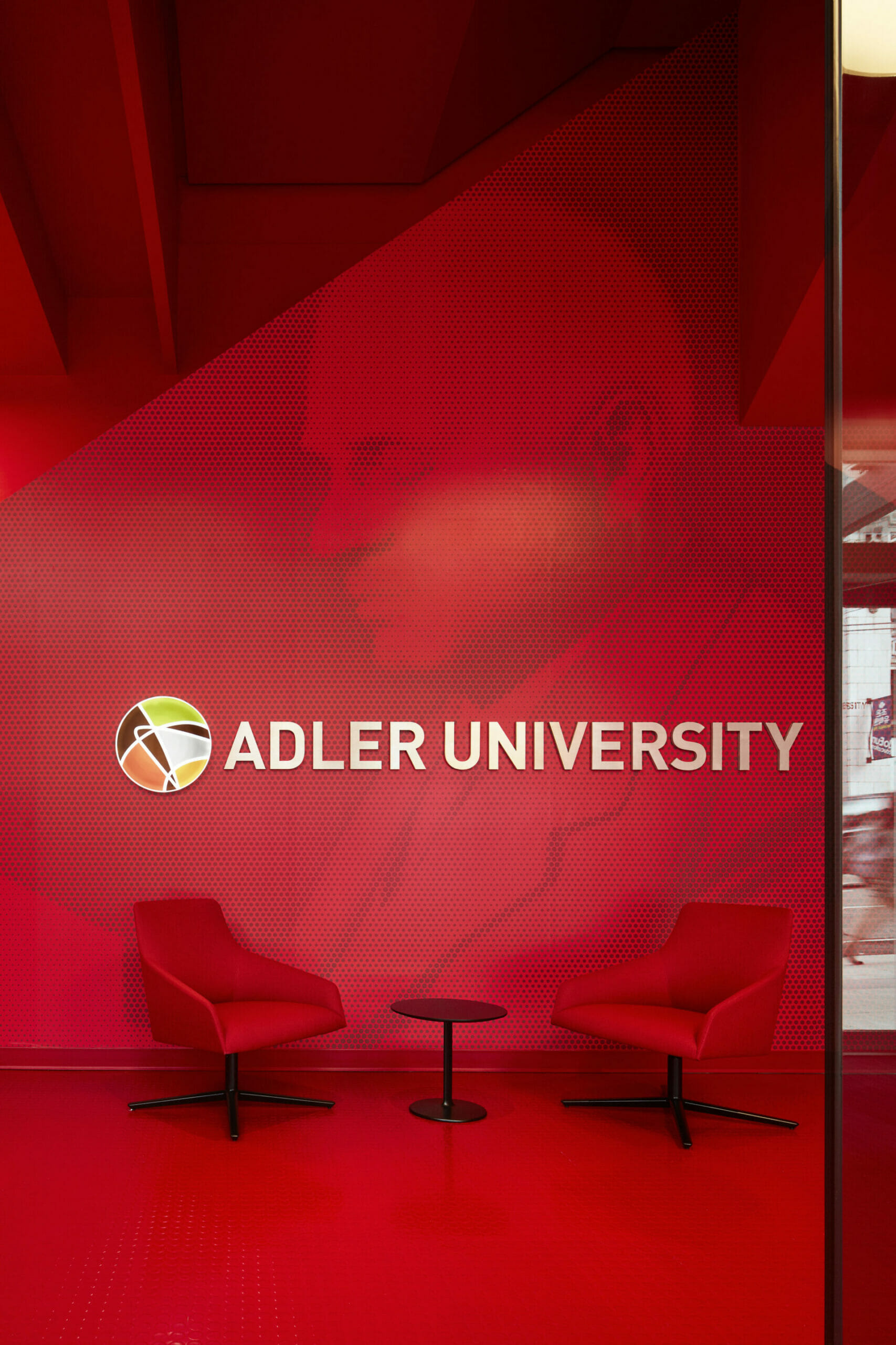 A red-toned lobby area with two chairs, a small table, and a wall featuring the Adler University logo and a large portrait in the background.