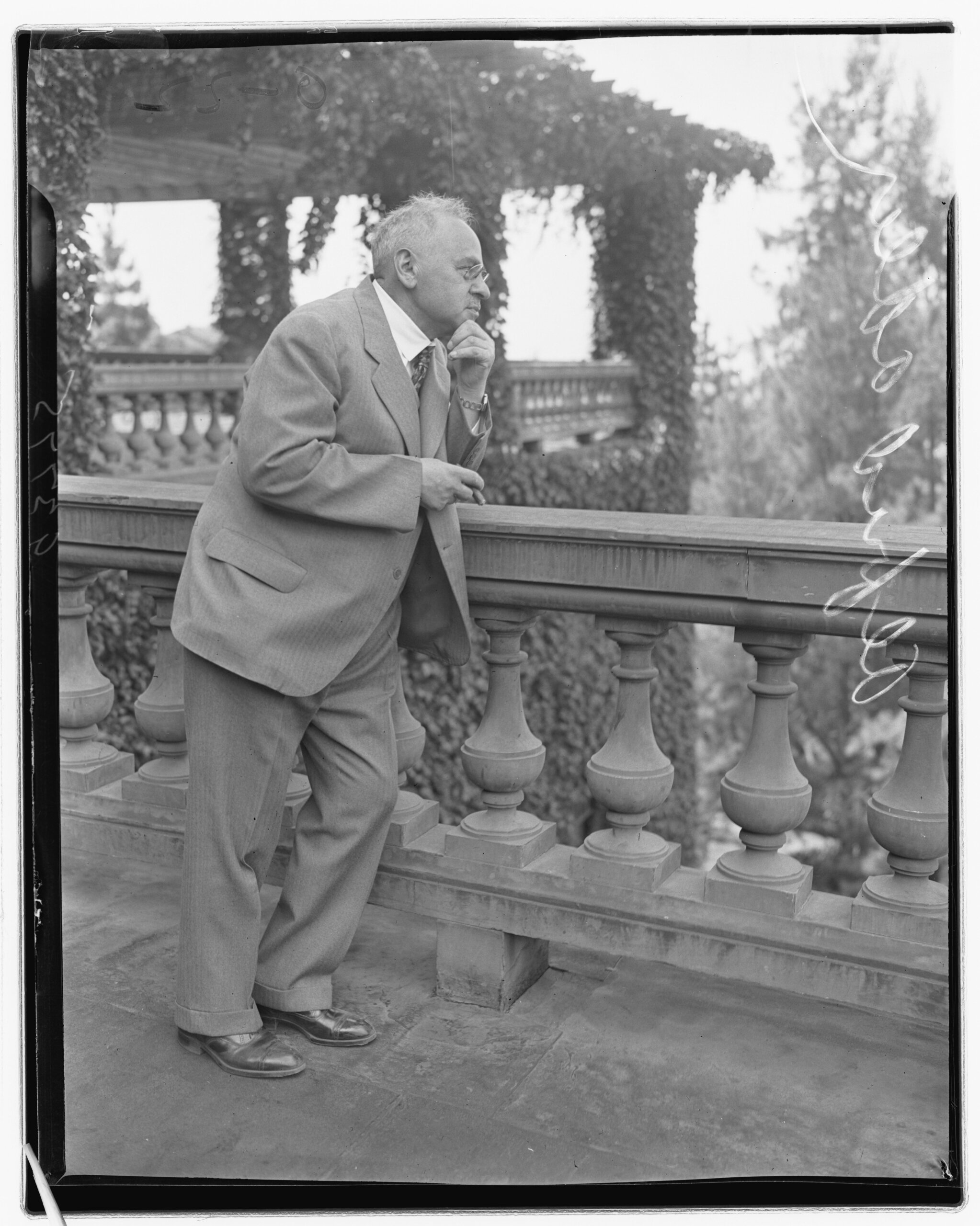 An older man in a suit, perhaps a psychologist inspired by Alfred Adler’s individual psychology, leans against a stone railing on a balcony, looking thoughtfully into the distance. Lush greenery surrounds the area.