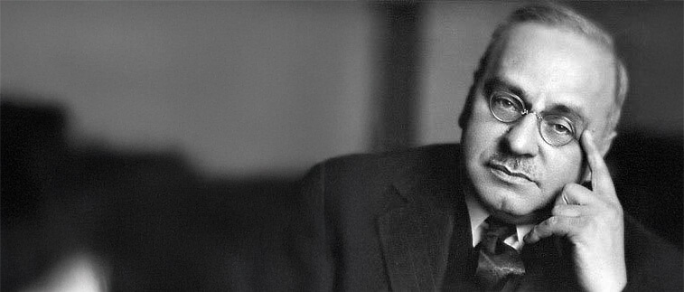 A black and white portrait of an older man with glasses, a mustache, and a suit, resting his head on his hand and looking at the camera.