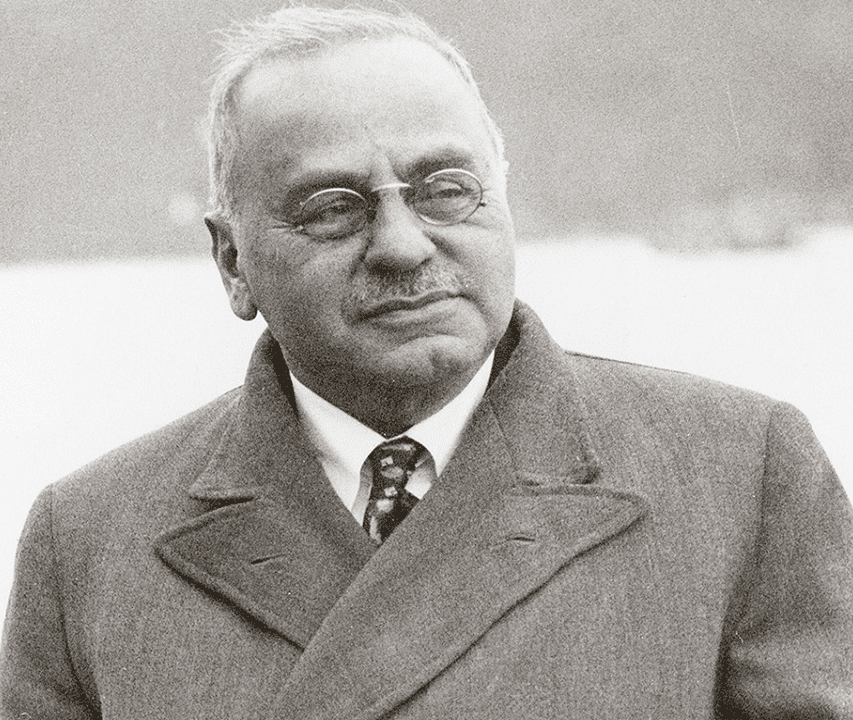 An older man with glasses and a mustache, wearing a coat and tie, looks slightly to the side in this black-and-white photograph.