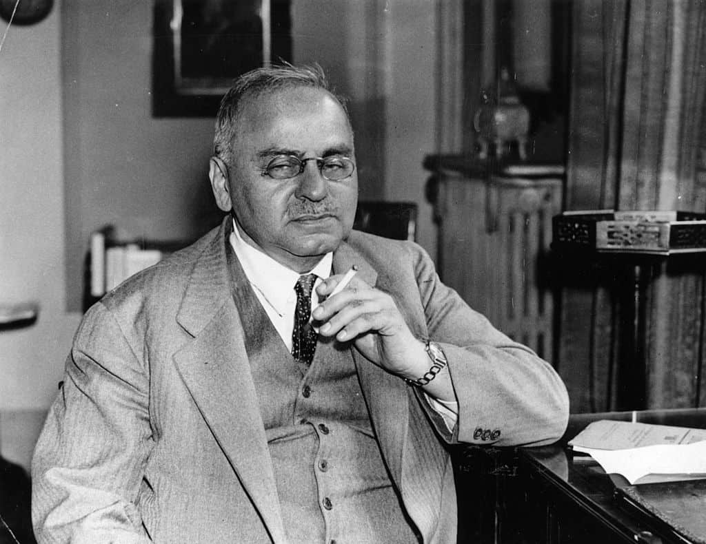 An older man in a suit sits at a desk, holding a cigarette and looking forward with a serious expression, reminiscent of Alfred Adler. Papers are visible on the desk.