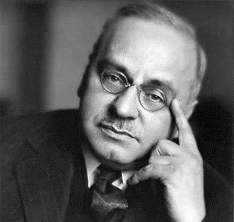 A man with glasses and a mustache rests his head on his hand, looking at the camera with a serious expression.