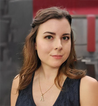 A woman with wavy brown hair, wearing a dark sleeveless top and a necklace with the letter "A," stands in front of a blurred, industrial background, ready to guide prospective students through available admissions and student resources.