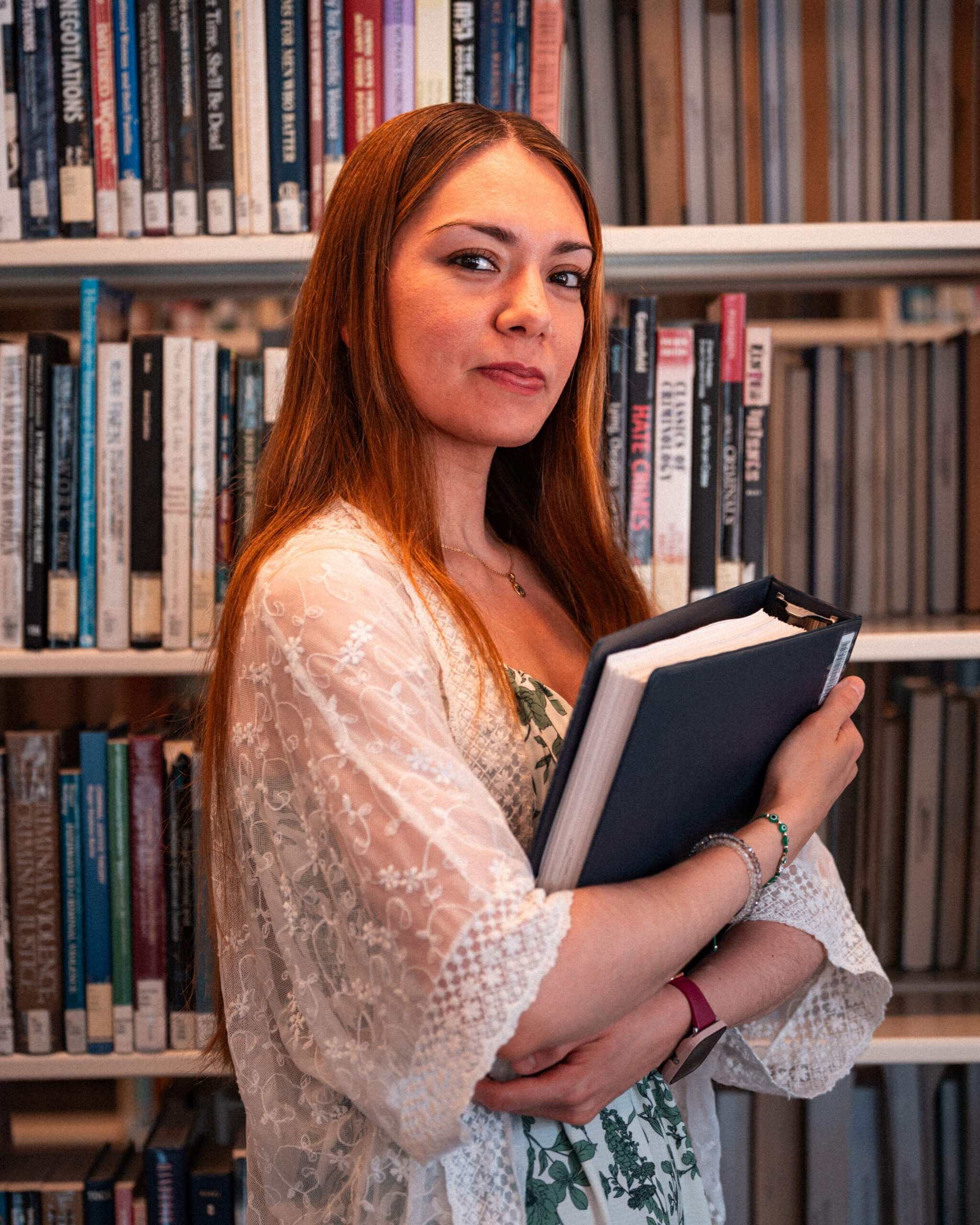 Woman with long auburn hair stands in front of bookshelves, holding books to her chest and looking at the camera. She is wearing a white lace shawl over a floral dress, embodying the strength found in alumni support and resources.