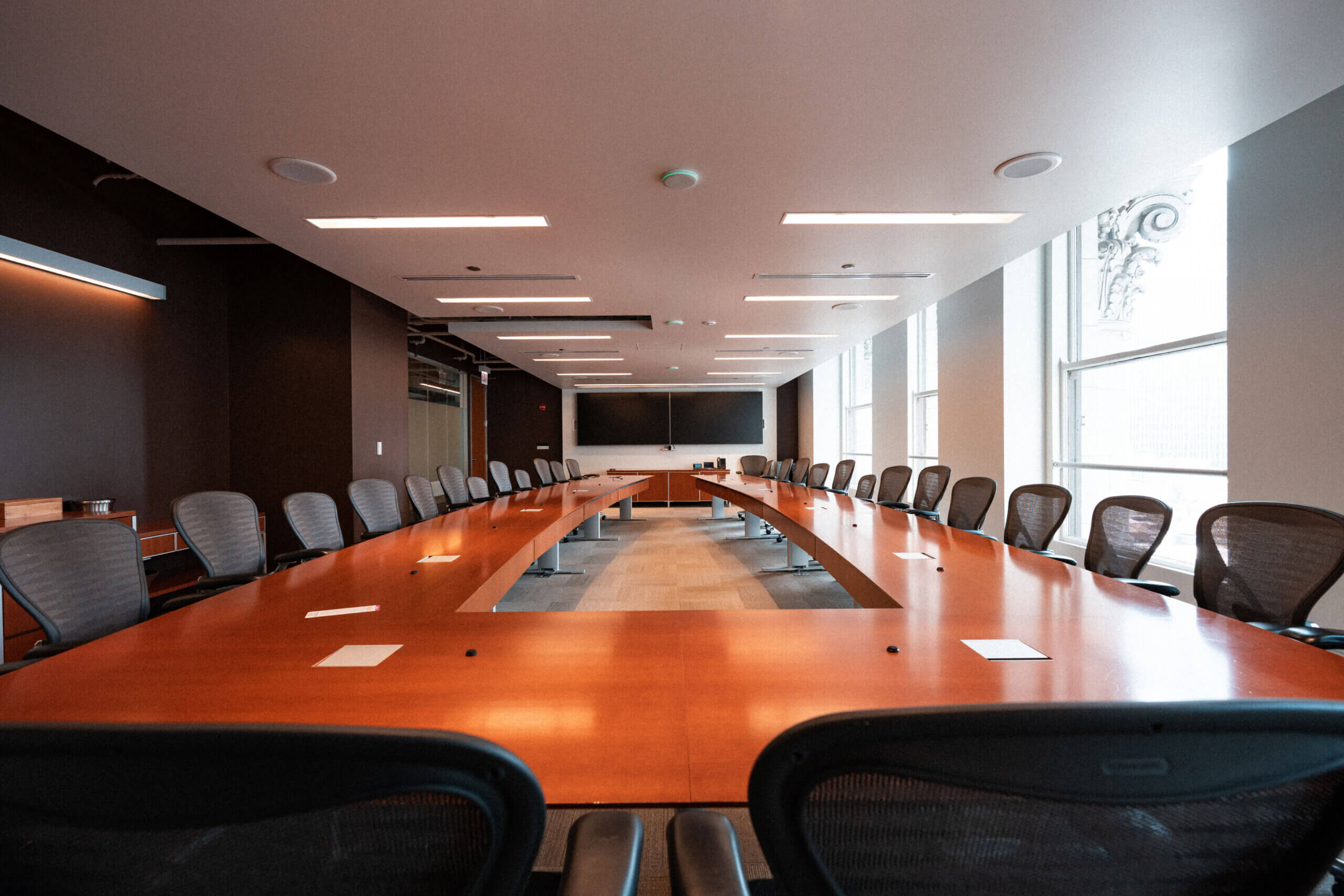 A large, empty boardroom with a long wooden conference table and black chairs awaits Donors, with large windows letting in natural light—an ideal space for planning donor support and organizing donation resources.