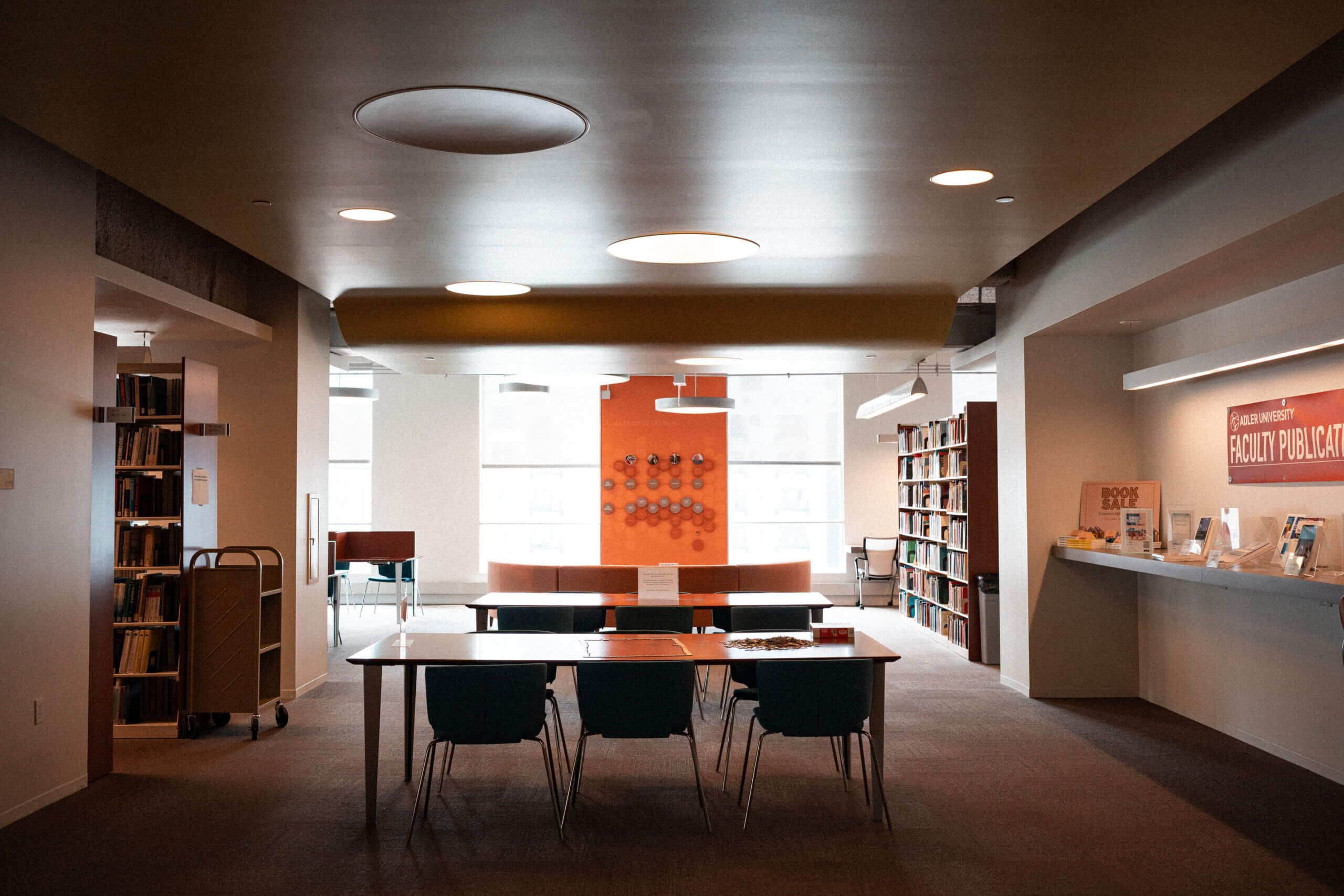 A modern library reading area with a table and chairs in the center, surrounded by bookshelves and a faculty publications display on the right, featuring donor information for those interested in supporting the collection.