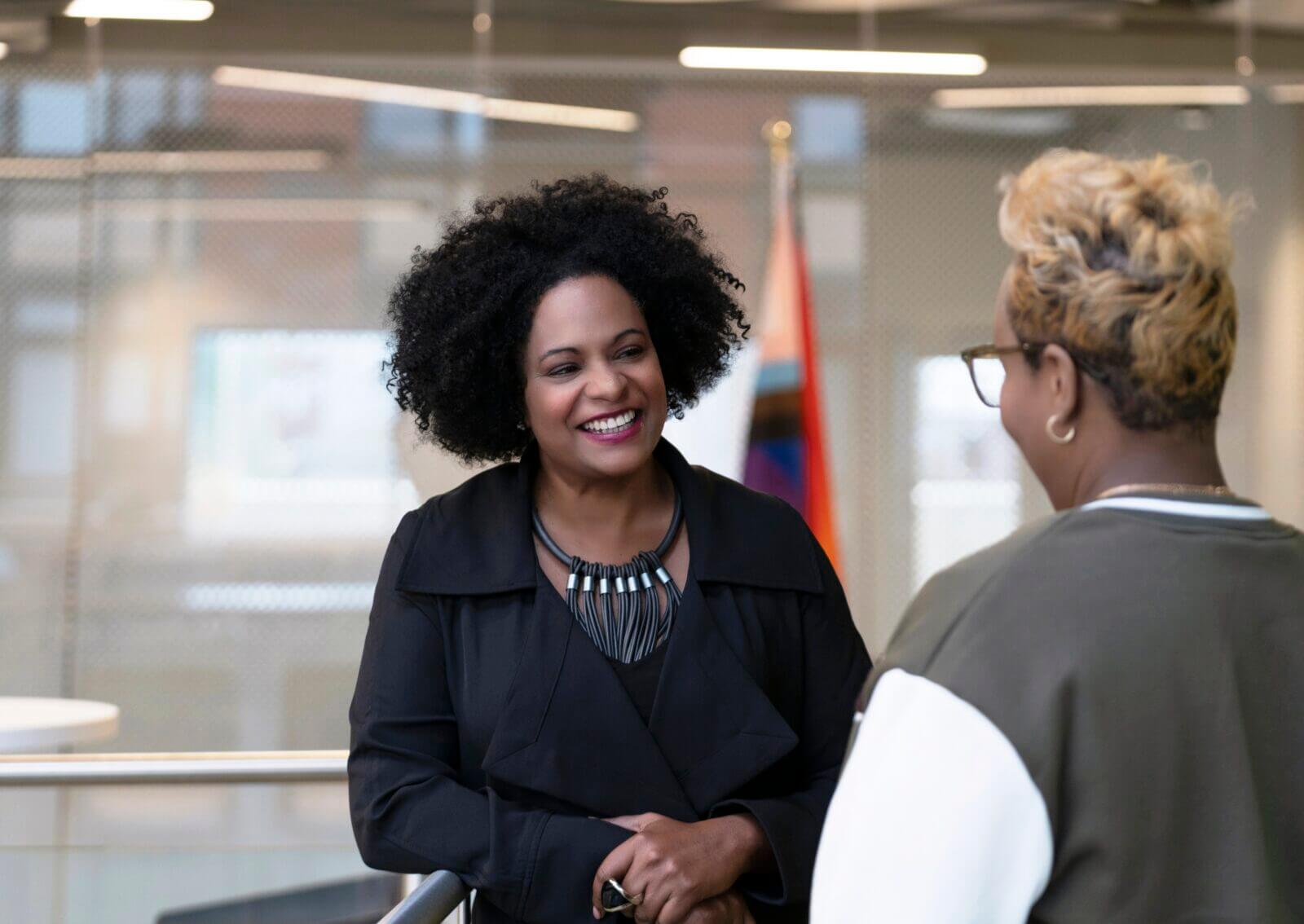 Two women are standing indoors, talking and smiling at each other, with a blurred flag in the background, perhaps discussing matters related to the President.
