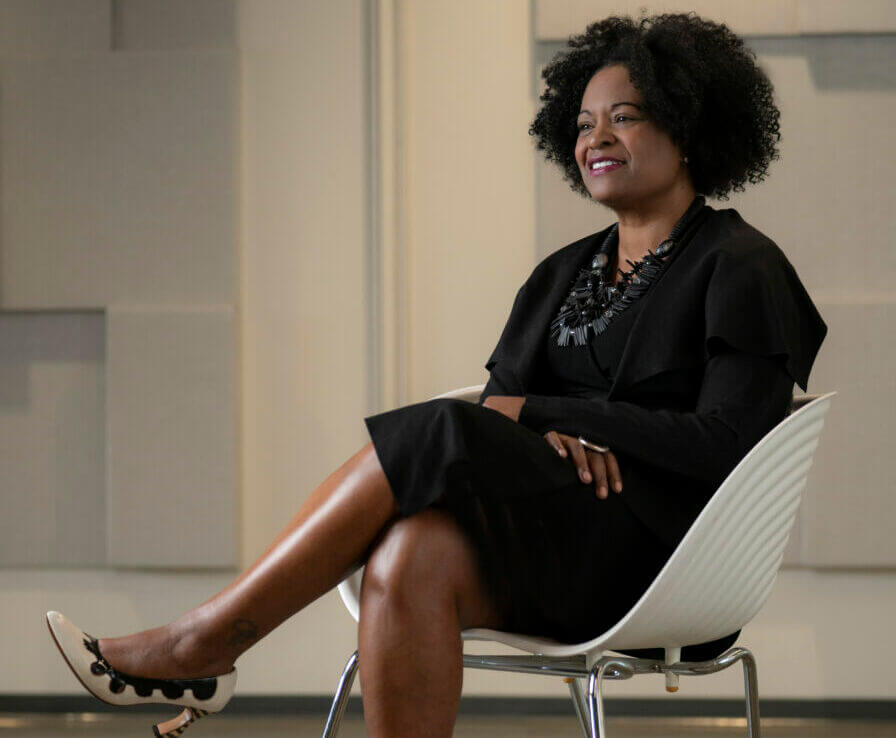 A woman with curly hair, resembling a president in poise, wearing a black dress and necklace, sits cross-legged in a white chair, smiling in a modern, neutral-colored room.