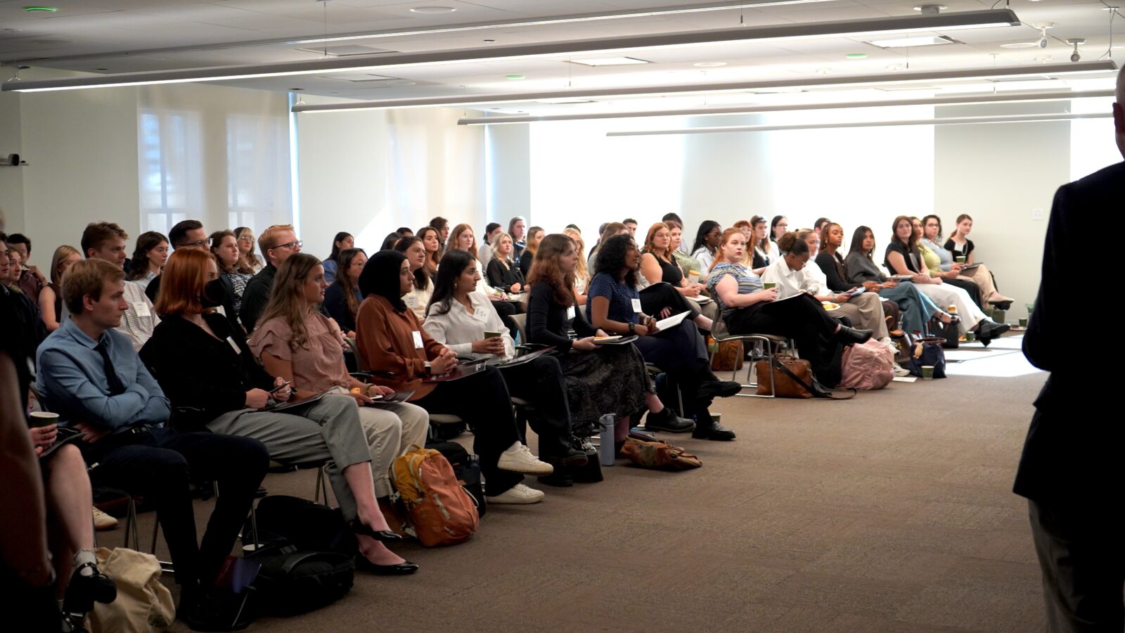 A large group of people sits in rows facing forward, attending an Erasmus+ presentation in a modern, well-lit conference room.