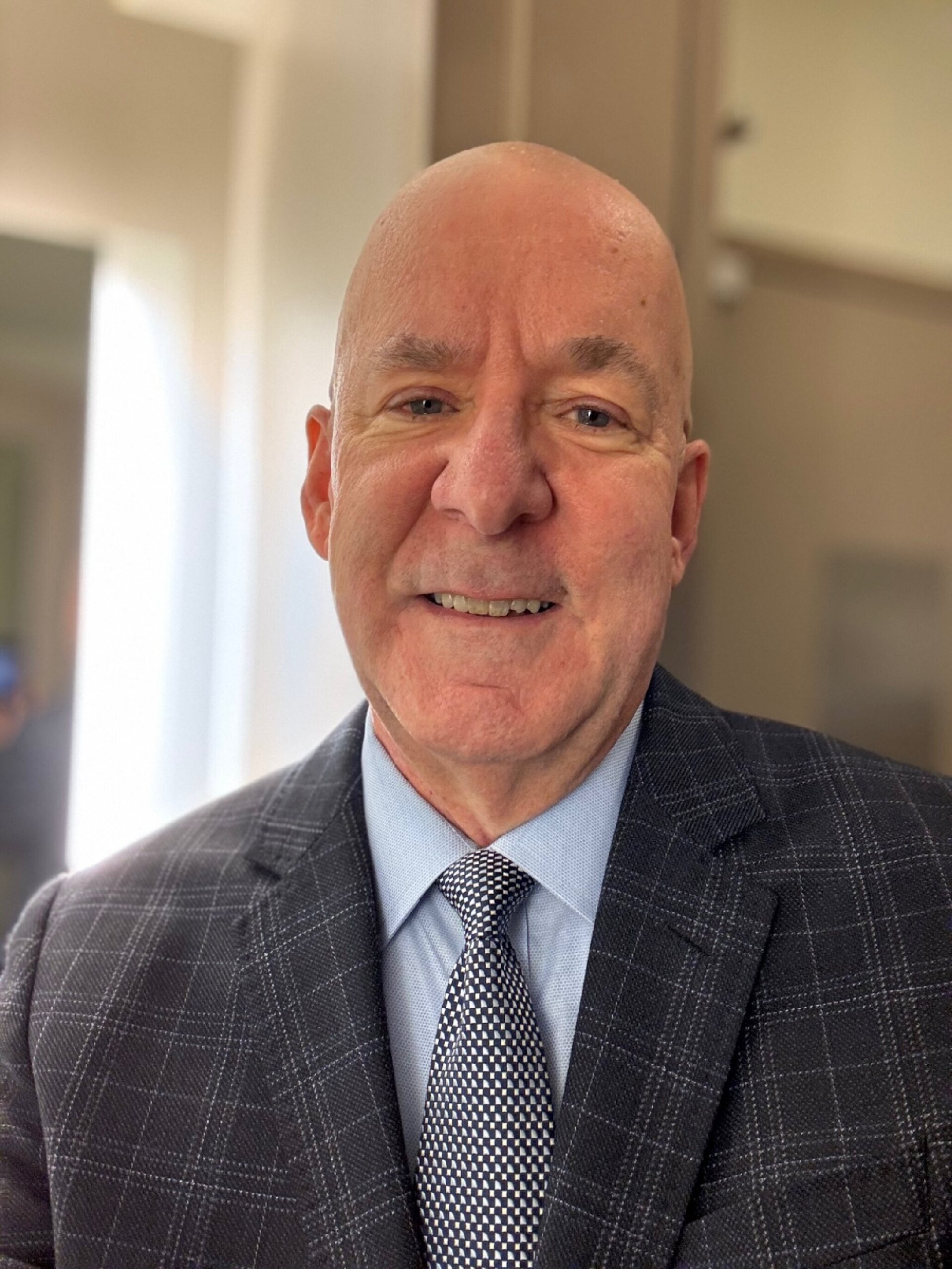 William Enhamm, a middle-aged man with a bald head, stands indoors in a gray plaid suit, blue shirt, and patterned tie, smiling slightly at the camera.