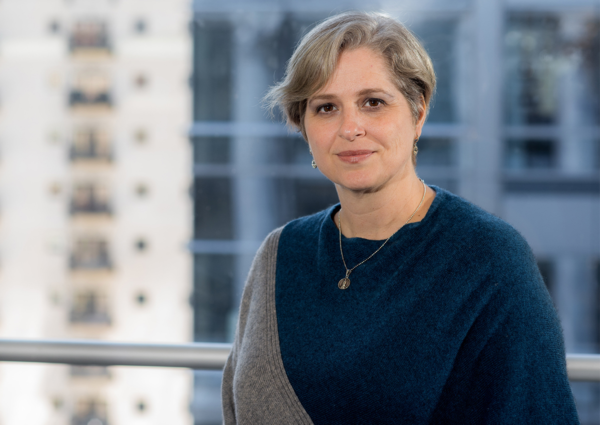 A person with short, light hair wearing a blue and gray sweater stands indoors with city buildings in the background—perhaps one of the dedicated scholarship applicants hoping for the Quintana Last Mile Scholarship.