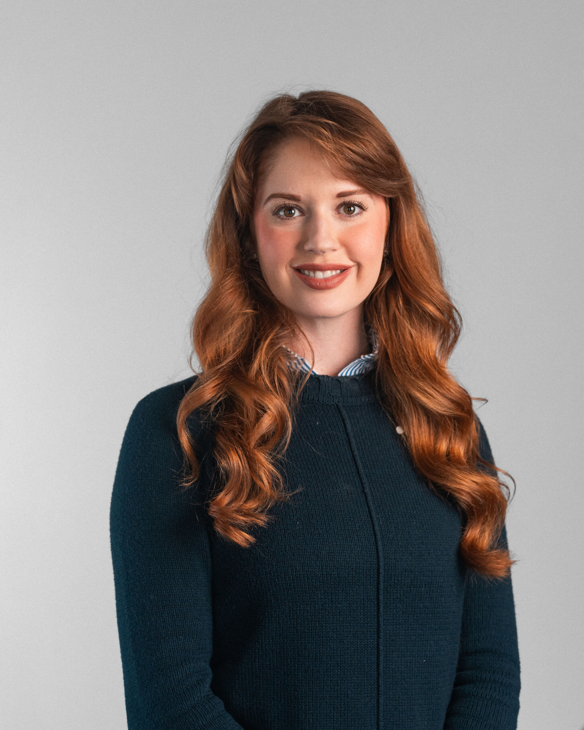 A woman with long, wavy red hair wearing a dark sweater stands against a plain light gray background, smiling at the camera with the confidence of senior leadership.
