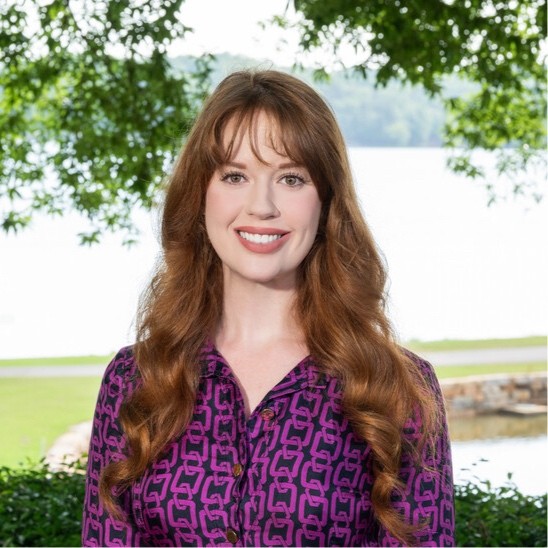 A woman with long red hair, wearing a purple patterned shirt, stands outdoors in front of a lake and trees, smiling at the camera, capturing the warmth of people and the beauty of nature in this vibrant scene.