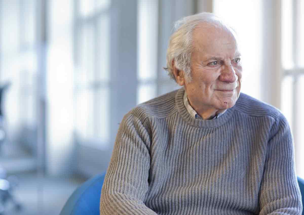 An older man with white hair and a grey sweater sits on a blue chair, looking to the side in a brightly lit room with large windows.