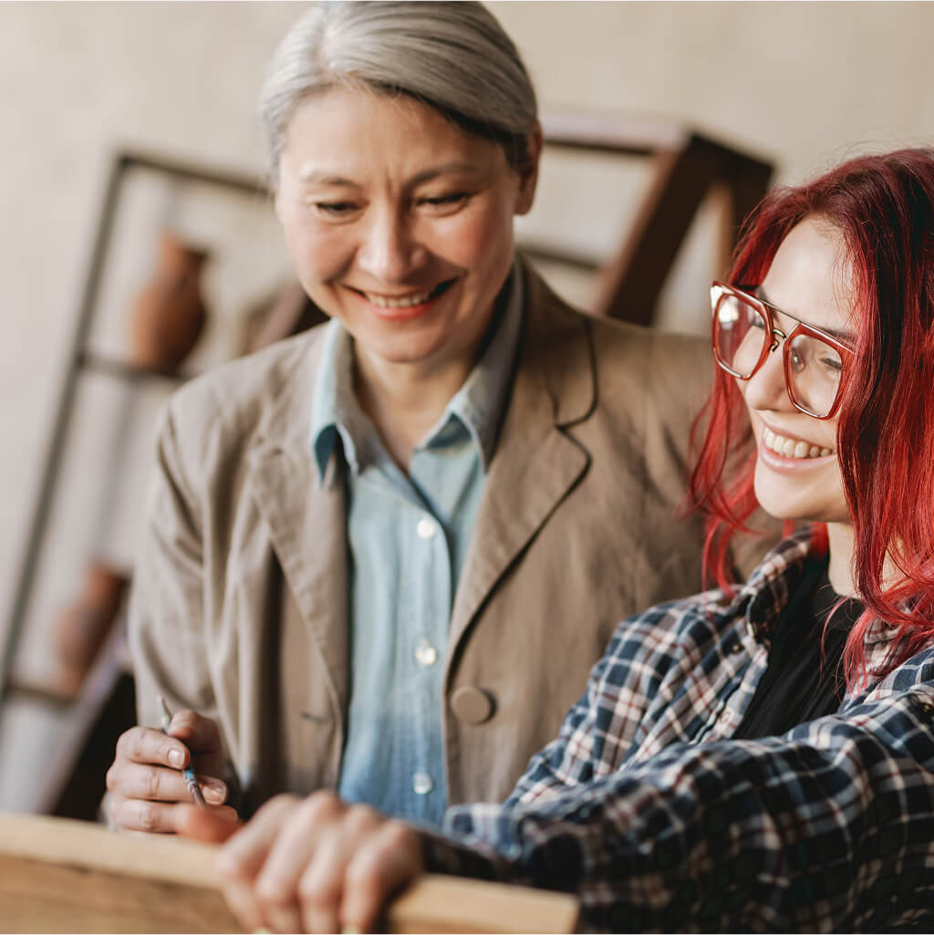Two women stand together indoors, one with gray hair and a beige jacket, the other with red hair and glasses—both smiling as they discuss M.A. studies in Art Therapy or Counselling Psychology.