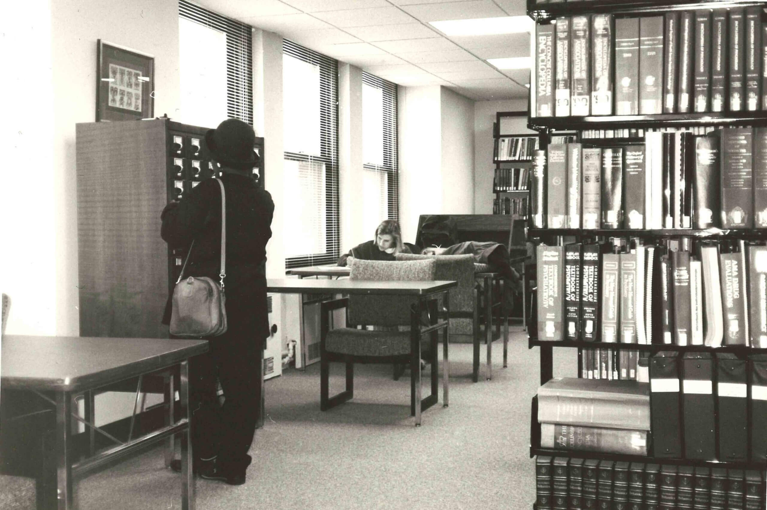 A person stands at a card catalog while another sits reading at a table in a library with bookshelves and large windows.