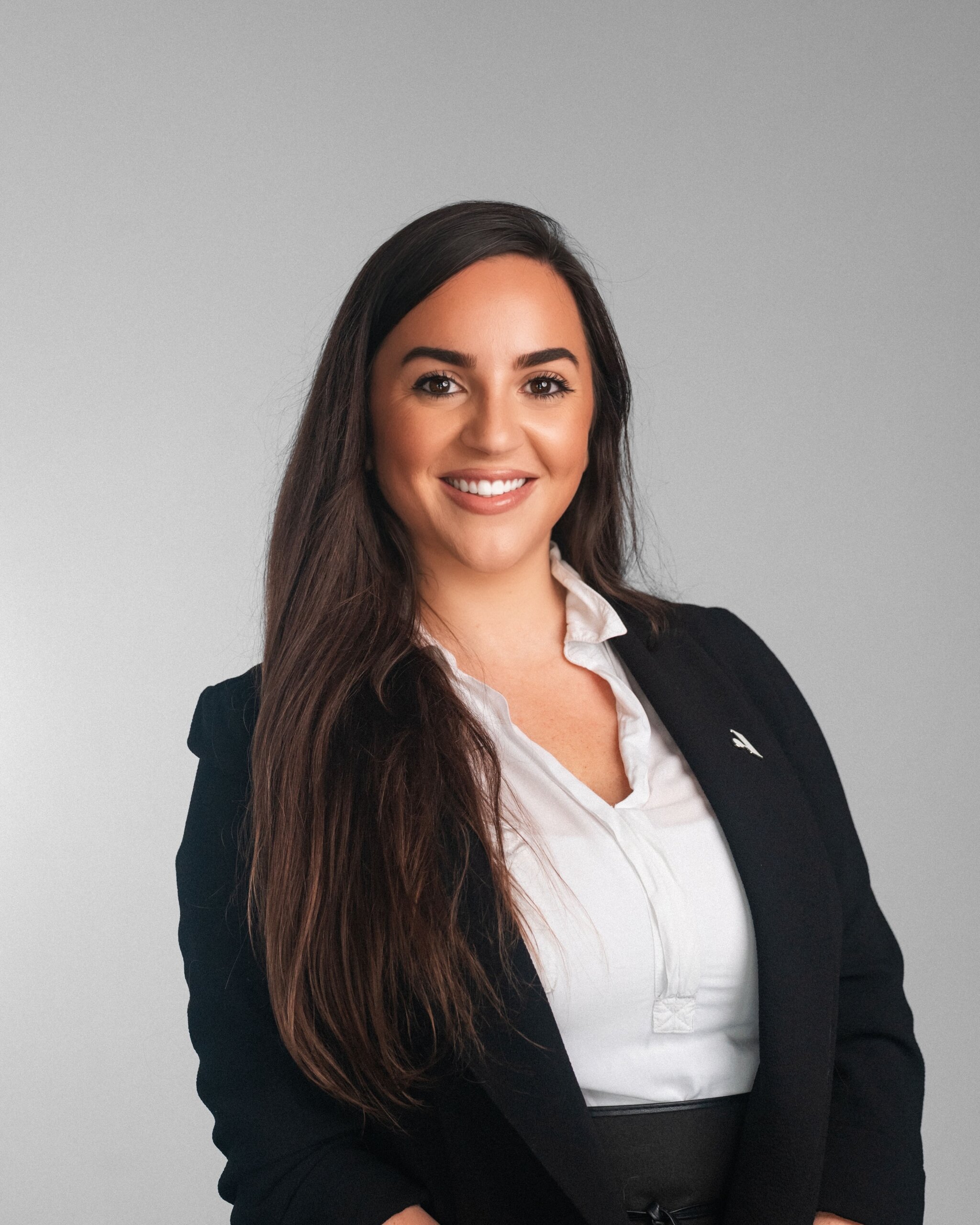 Tara Romanella, a woman with long brown hair in a black blazer and white blouse, smiles warmly in front of a plain gray background.