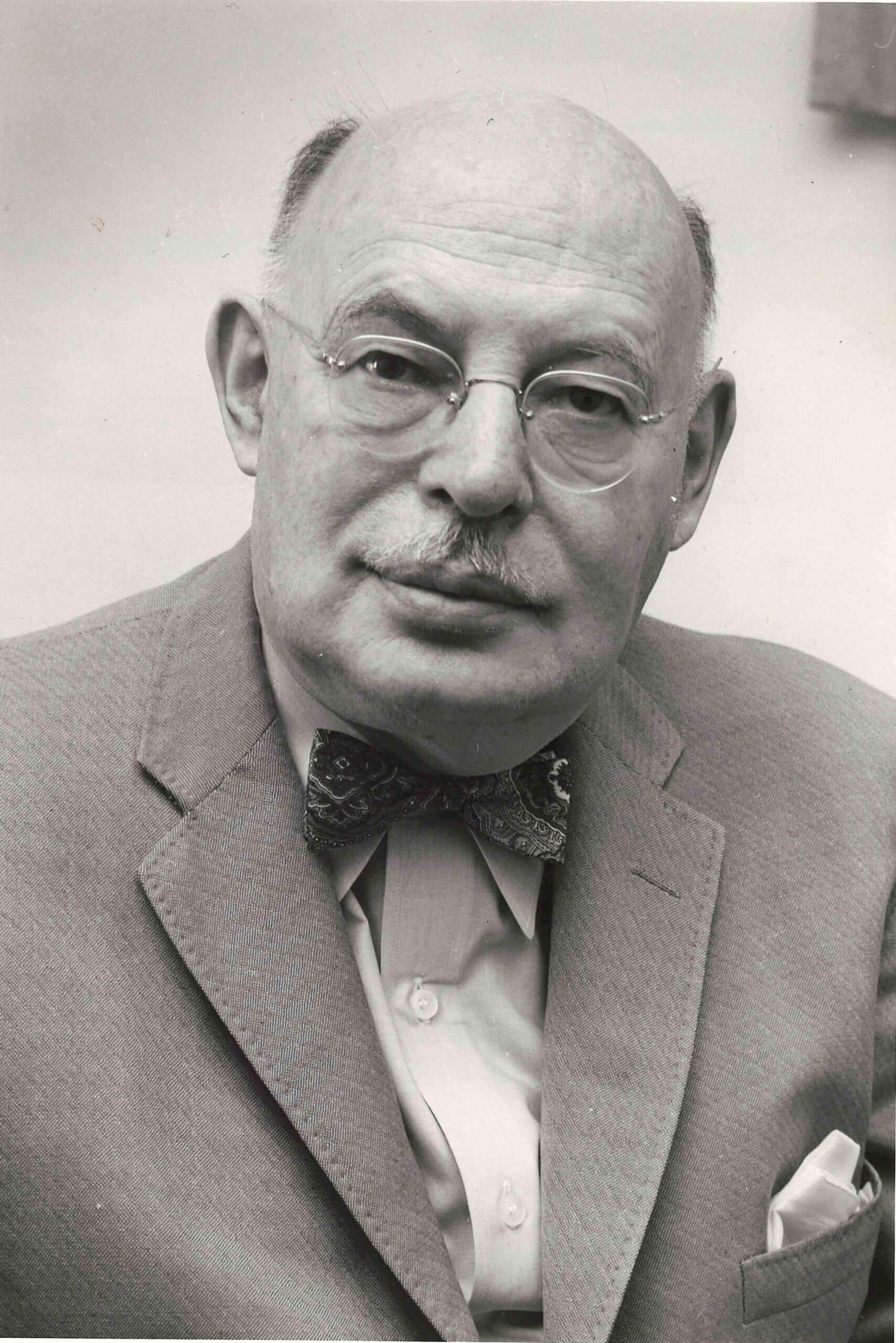 An older man with glasses, a mustache, and a bow tie, wearing a suit jacket and collared shirt, poses for a black and white portrait.