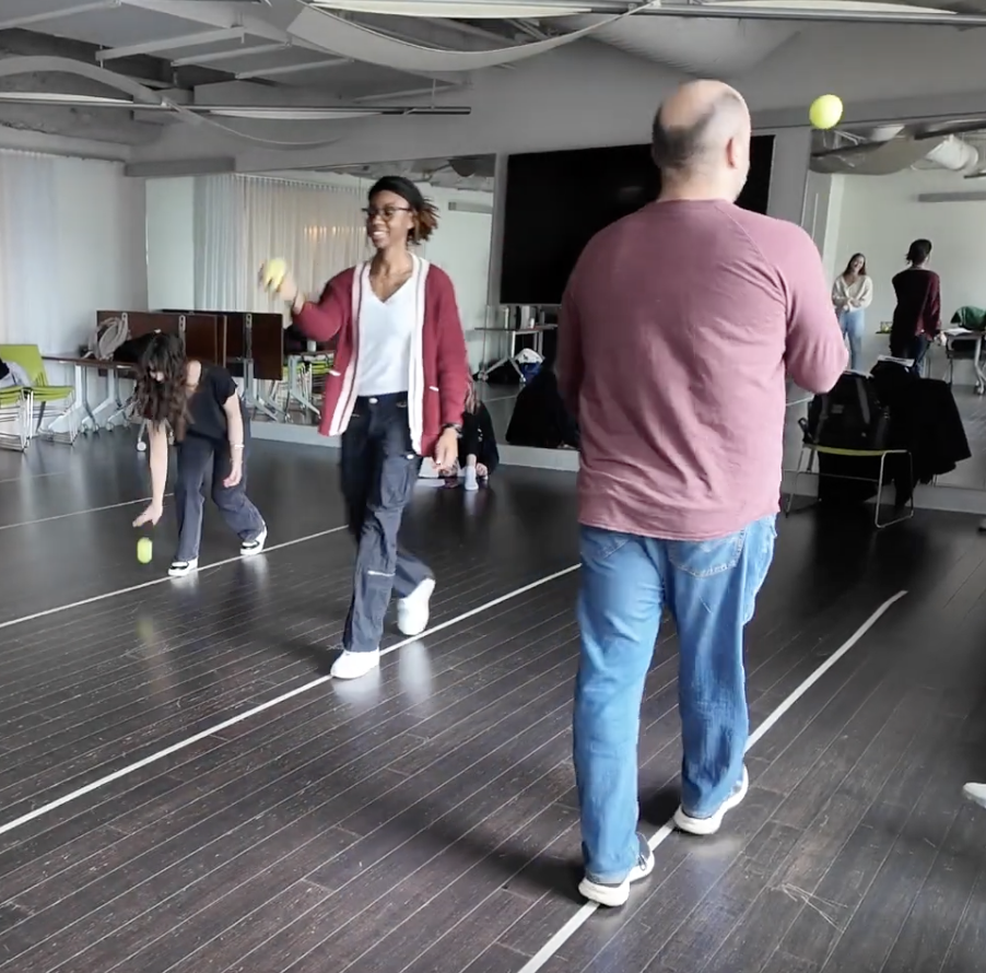 Three people in a room with wooden floors are tossing yellow balls, with chairs and a large TV in the background.