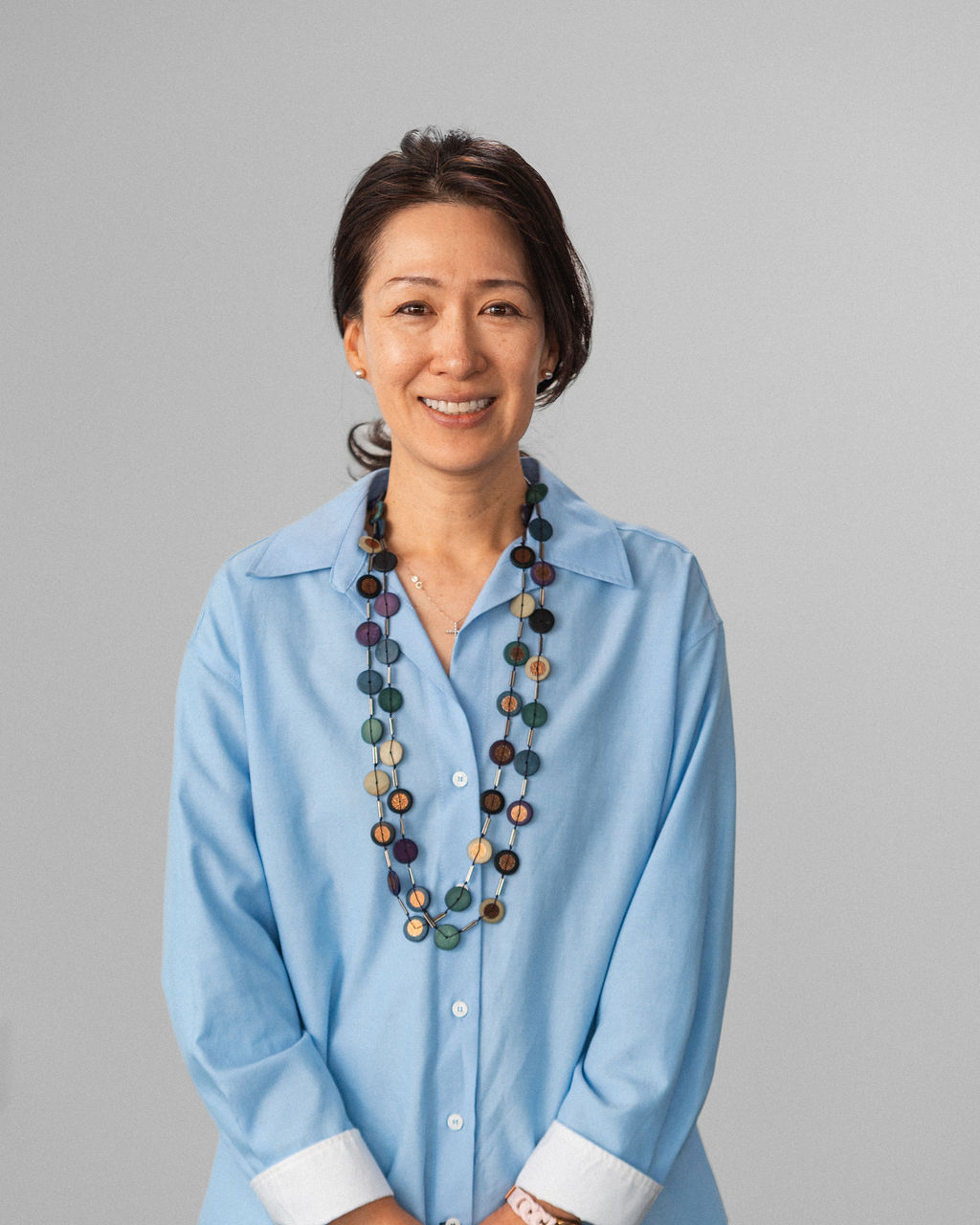 Jinnie, a woman with dark hair wearing a light blue collared shirt and a colorful beaded necklace, stands against a plain gray background, smiling at the camera.