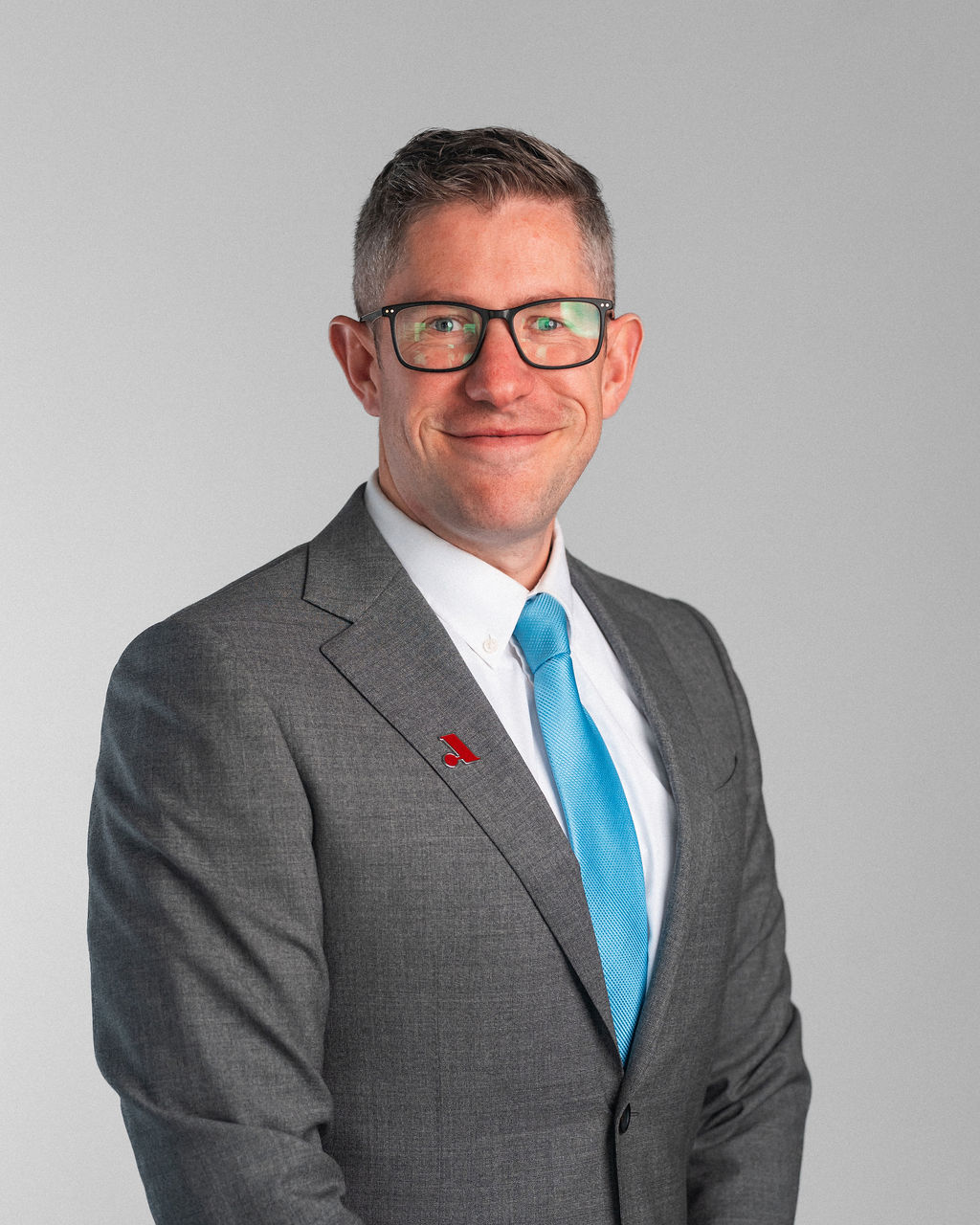 A man wearing glasses, a gray suit, white shirt, and blue tie stands against a plain light gray background, smiling at the camera.