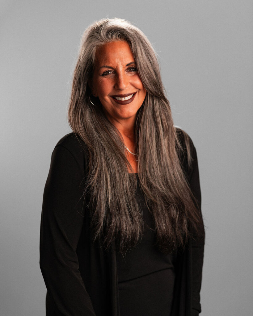 Jennifer La Civita, a woman with long gray hair, wearing a black top and blazer, stands smiling in front of a plain gray background.