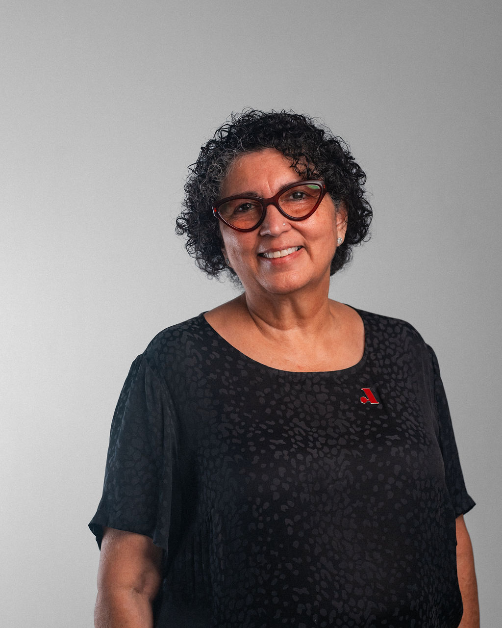 Rita Dibble, a woman with short curly hair and glasses, is smiling while wearing a black blouse and a red ribbon pin, standing against a plain light gray background.