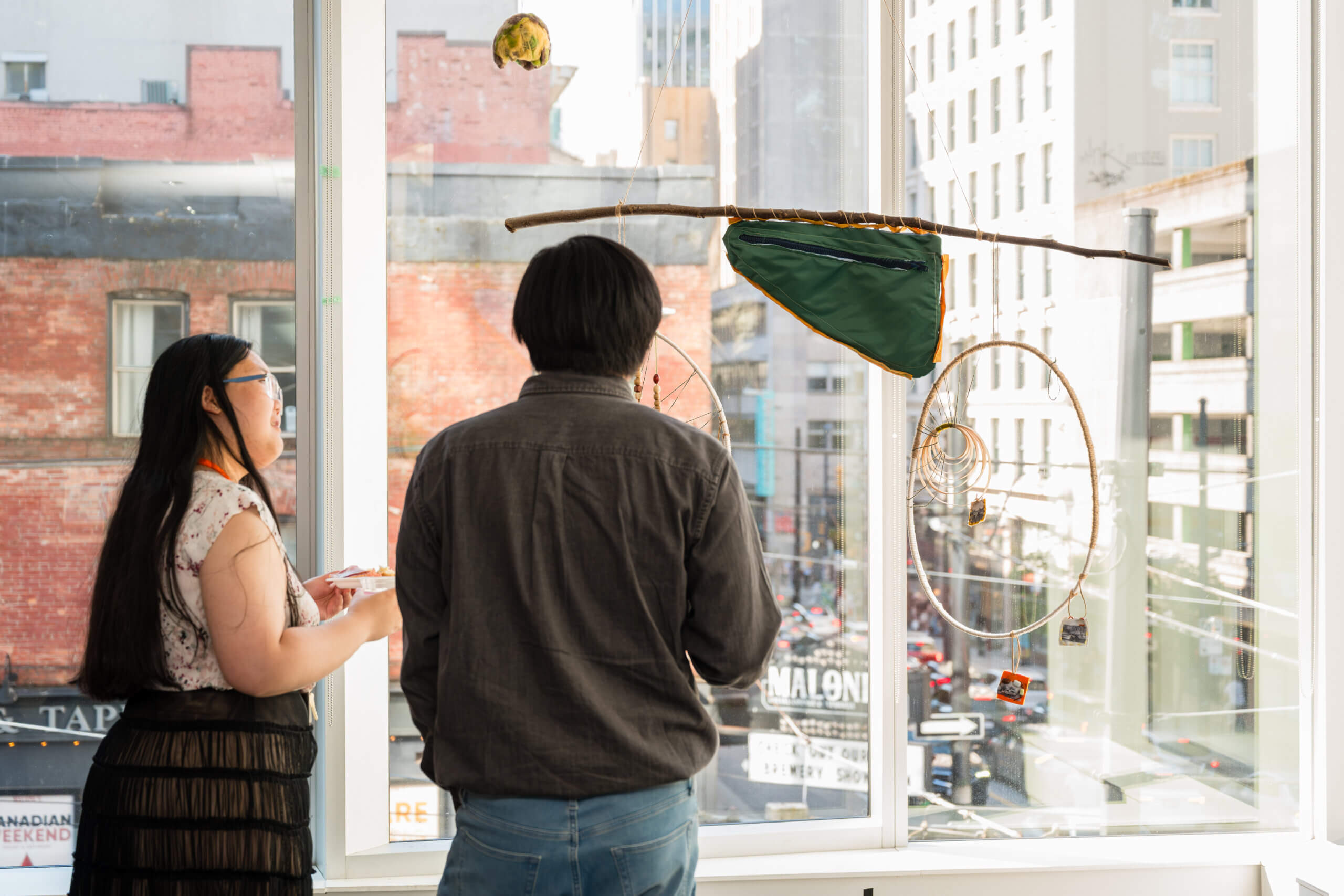 Two people stand by a large window looking at a hanging art piece made of sticks, fabric, and circular wire shapes, with a city street visible outside and donor resources available to support the arts.