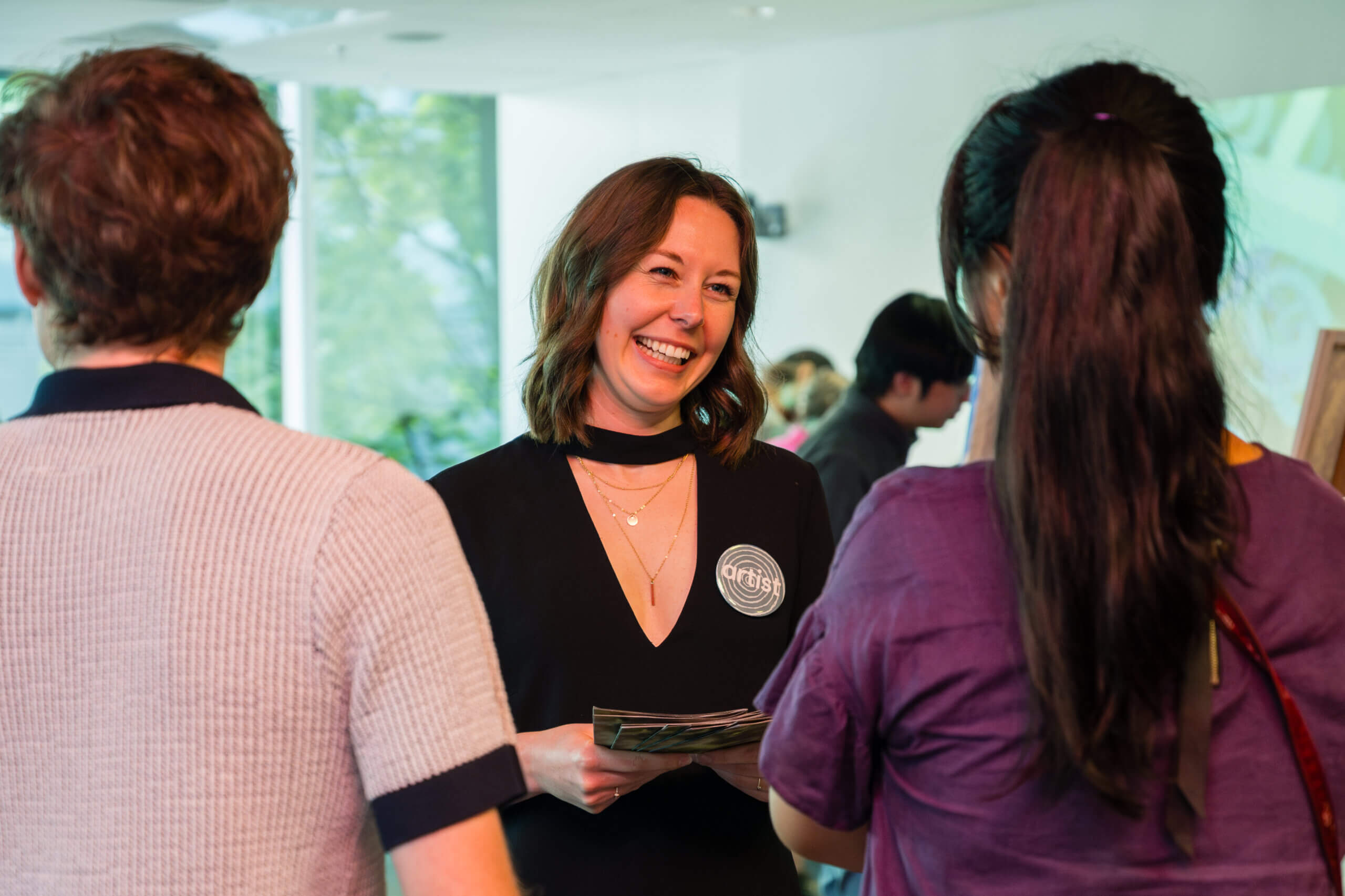 A woman smiling and talking with two people at an indoor event. She holds papers and wears a badge on her black dress. The background shows more people and large windows with greenery outside.