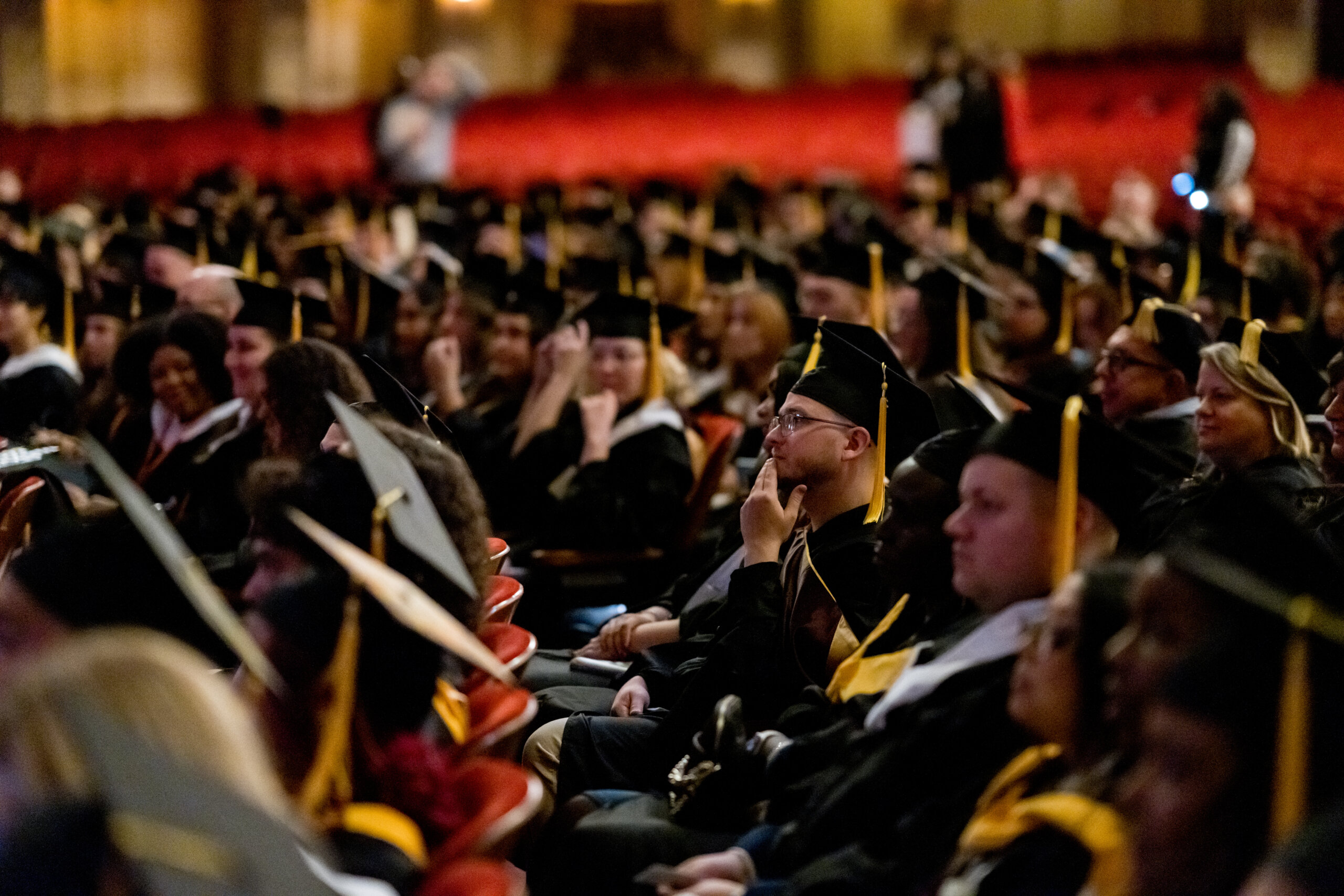 A large group of graduates in caps and gowns sit in rows of seats at an indoor ceremony, facing the stage, ready to join a network of alumni resources.