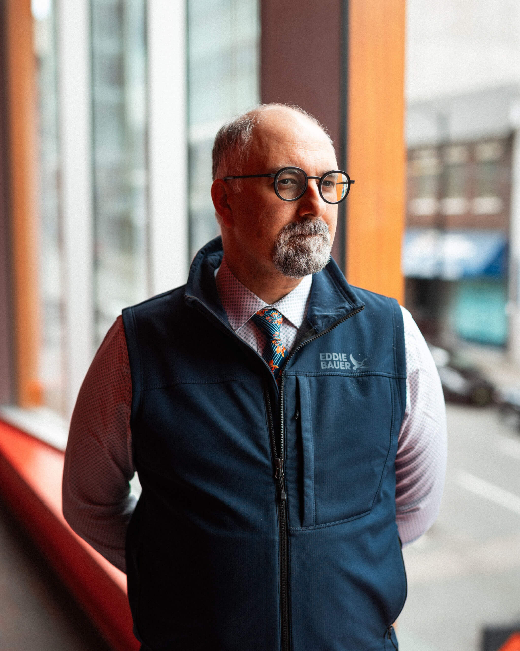 A man with glasses, a beard, and a mustache wearing a blue Eddie Bauer vest and patterned tie stands indoors next to a window, looking outside—perhaps reflecting on opportunities for prospective students or student resources available.