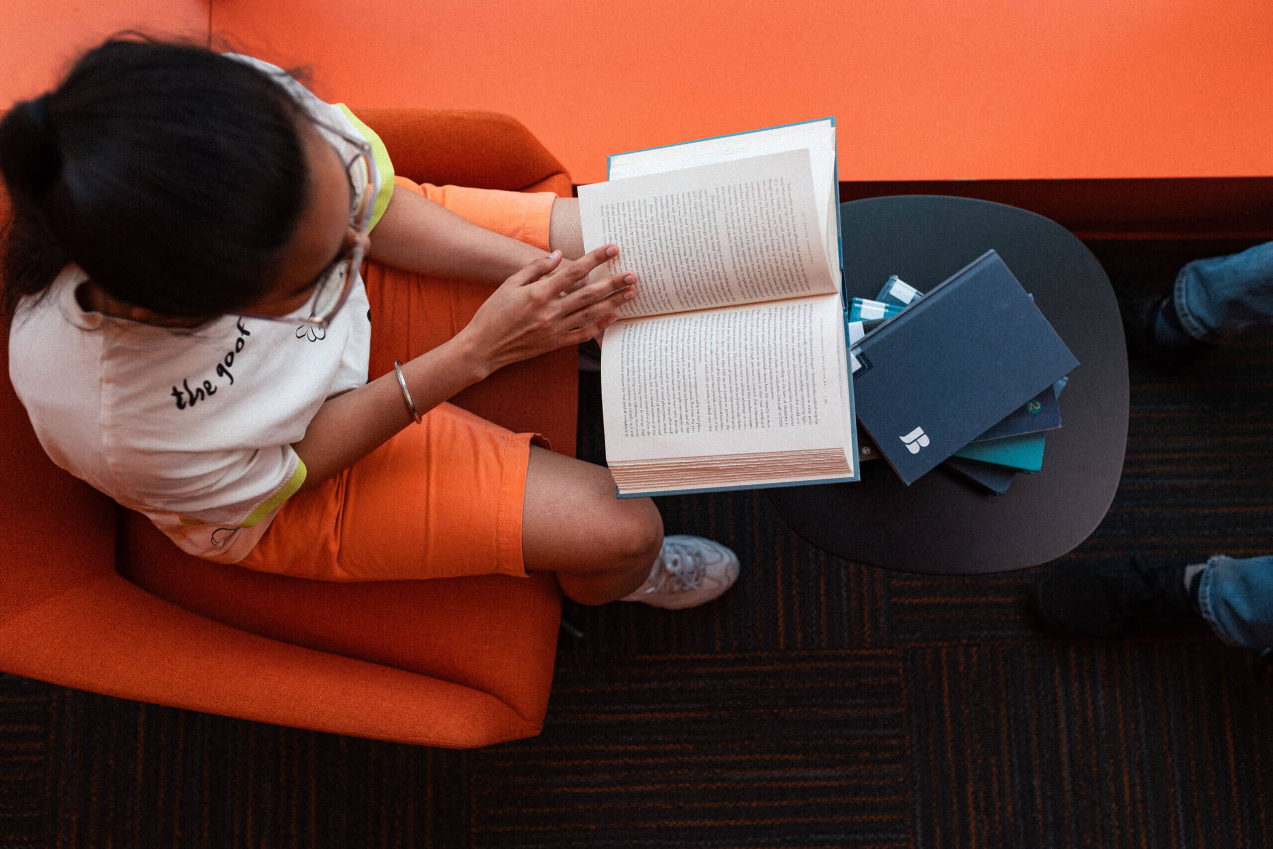 A person sitting in an orange chair reads an open book about donor resources, with more books and notebooks on a small black table beside them.