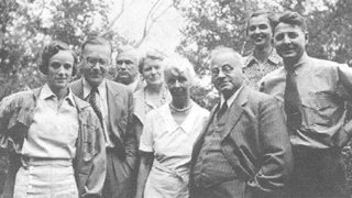 A group of seven adults, four men and three women, pose outdoors for a black-and-white photograph. Among them is Alfred Adler, with trees visible in the background.