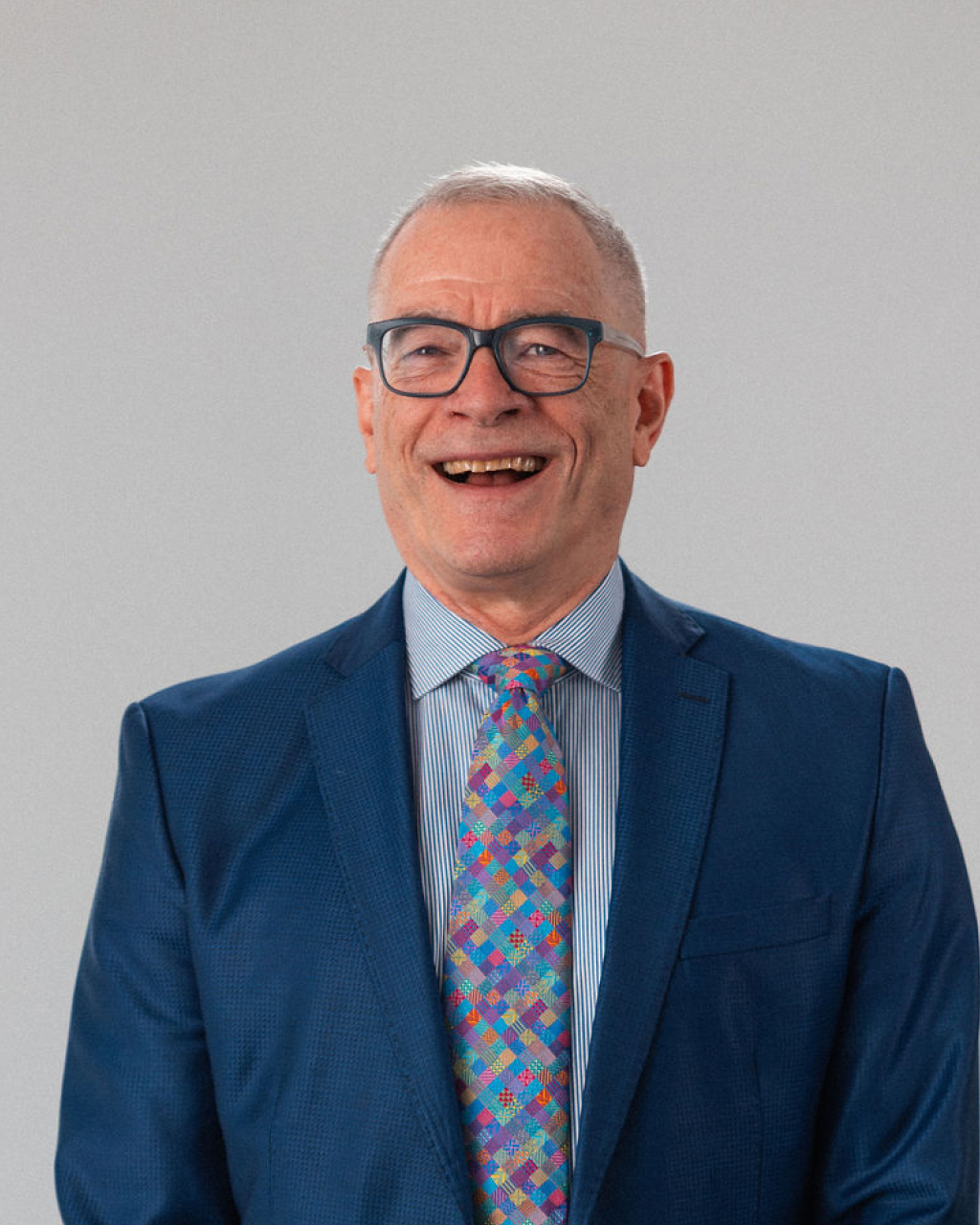 A middle-aged man in a blue suit, striped shirt, and colorful patterned tie, smiling and standing against a plain light background.