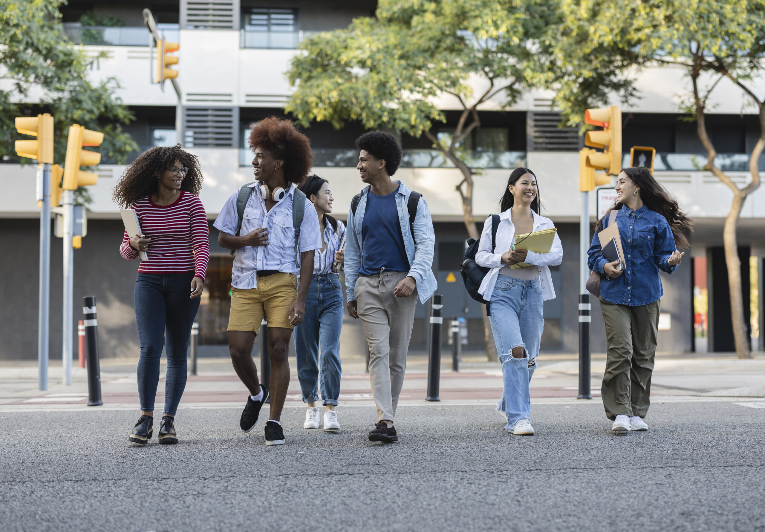 Six young adults with backpacks and books walk together across a street in an urban area, talking and smiling, as trees and an institute building focused on public safety rise in the background.