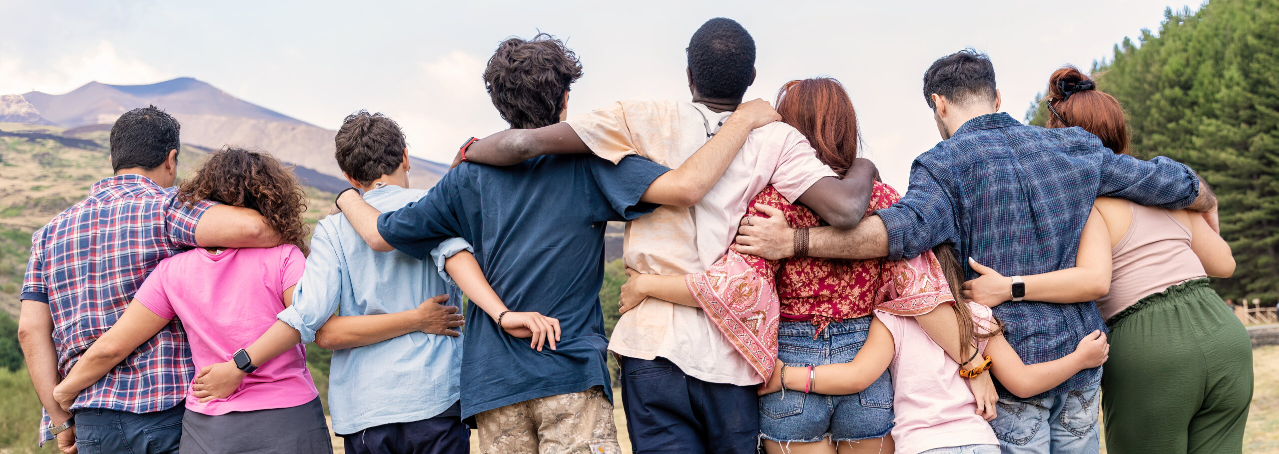 A group of people stand outdoors with their backs to the camera, arms around each other, symbolizing unity and commitment to social justice as they face a breathtaking mountainous landscape and trees.