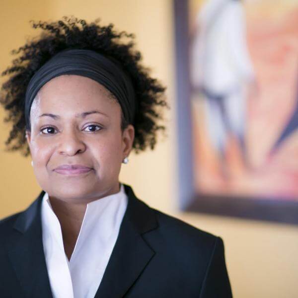 A woman with short curly hair and a headband, dressed in a black blazer and white shirt, stands indoors—with the confidence of a President—framed by a blurred painting in the background.