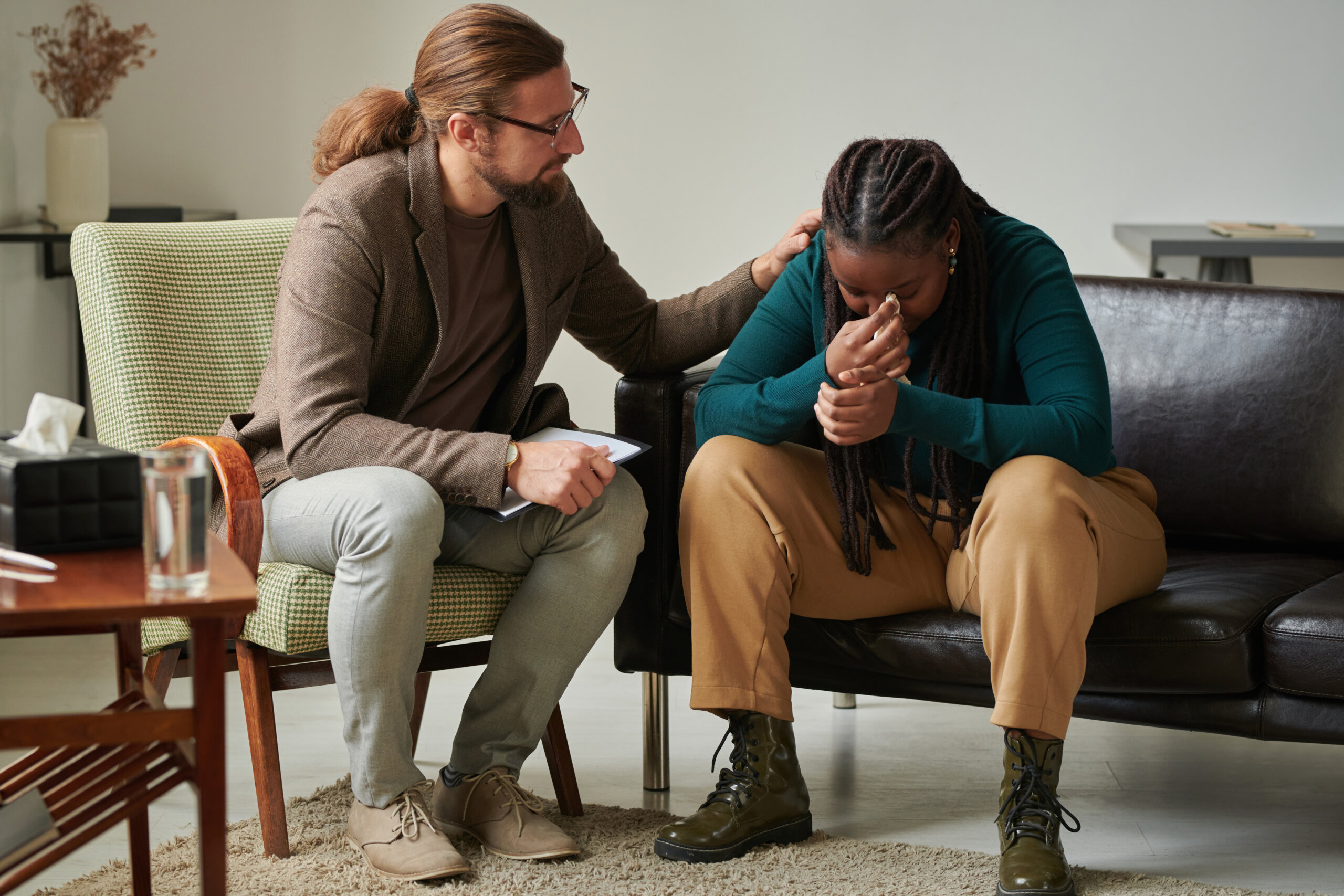 A man sits beside a woman who is visibly upset, comforting her with a hand on her shoulder in a counseling session focused on social justice.