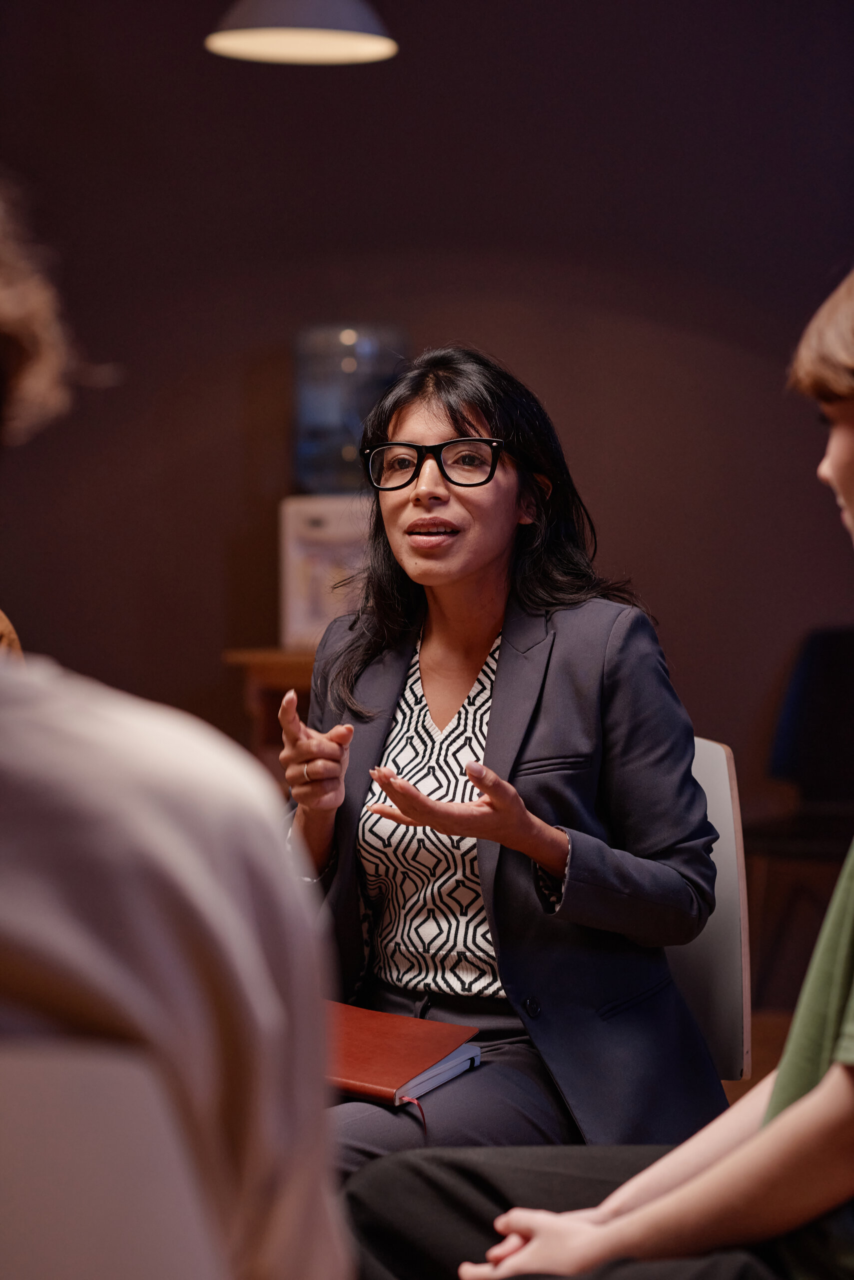 A woman in a suit and glasses speaks to two people in a dimly lit room, gesturing with her hands as they discuss issues of public safety.