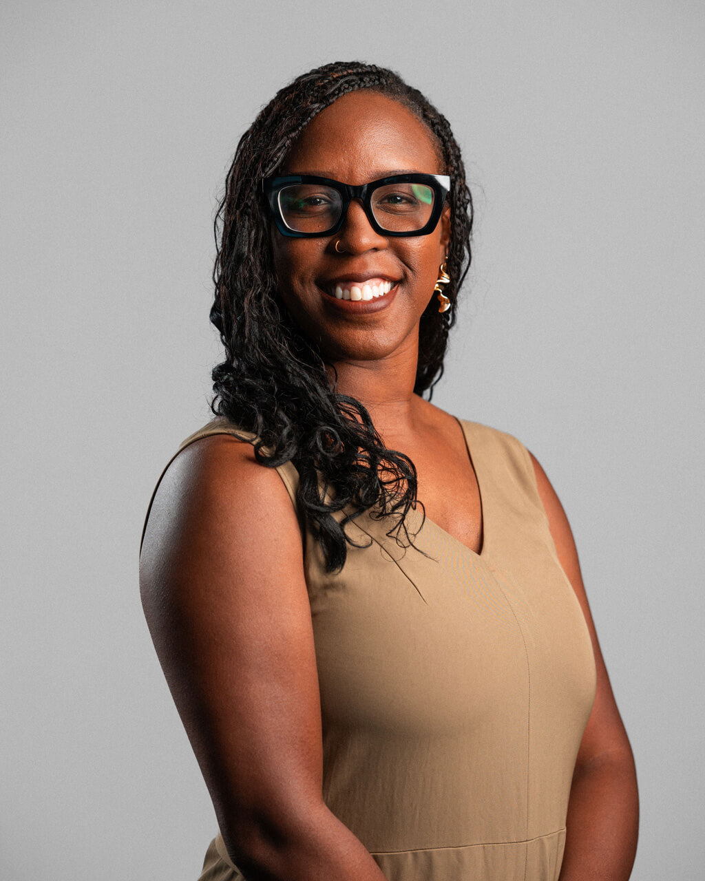 Dara Winley, a woman with glasses and long braided hair, smiles while standing against a plain light gray background. She is wearing a sleeveless beige dress and gold earrings.