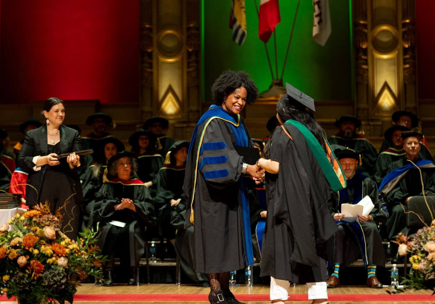 A graduate in cap and gown shakes hands with a faculty member on stage during an Adler University Commencement ceremony. Other faculty and graduates are seated in the background.